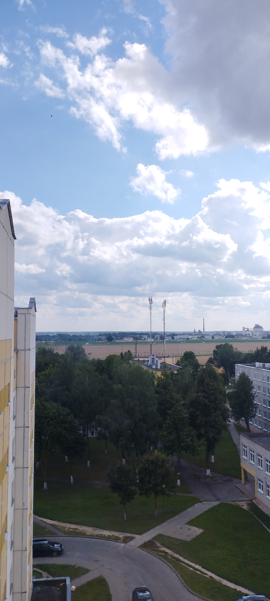 A vertical, eye-level shot captures a cloudy, bright sky above a cityscape. The left side of the frame is dominated by the side of a tall, light-colored apartment building with cream and yellow accents. Below the sky, a dense line of dark green trees stretches across the mid-ground, with some lighter green trees interspersed. Behind the trees, the landscape opens up to fields of light brown and yellow, suggesting agricultural land. Two tall, thin communication towers, each with multiple antennas, stand prominently in the distance. Further in the background, industrial structures with chimneys and silo-like buildings are visible. A grey apartment building with rows of white-framed windows is situated on the right side of the frame. The foreground shows a winding asphalt road and a green grassy area with scattered trees that have white trunks. Two dark-colored cars are parked on the road.