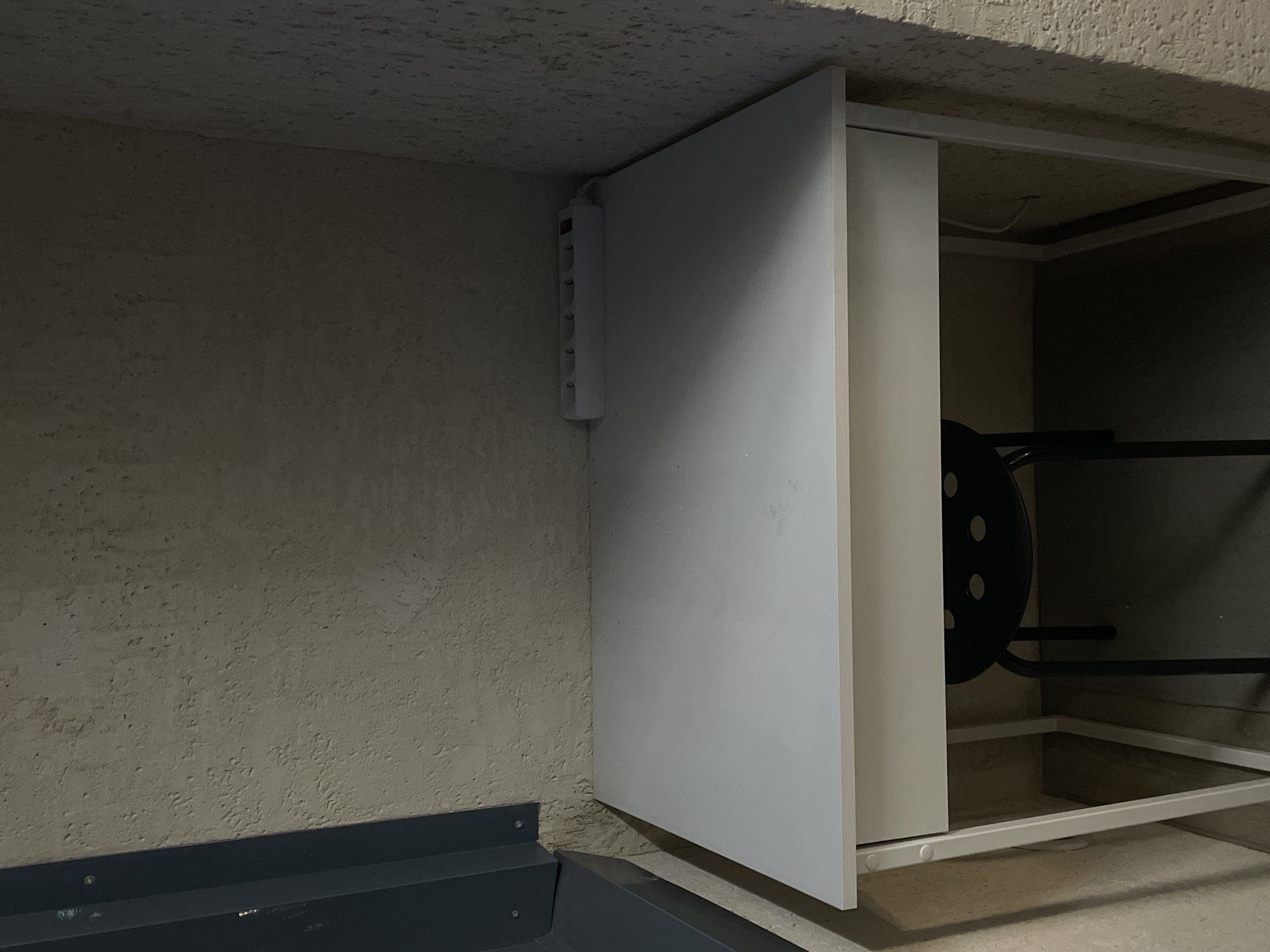 A white desk with a drawer is positioned in a narrow alcove. On the desk, a white power strip with six outlets and an on/off switch is located towards the back right. A black stool with a circular seat is placed underneath the desk. The walls of the alcove are textured and light-colored. To the left of the desk, a dark grey, angular object is partially visible. The scene appears to be indoors, in a space that could be an office or study area. The overall lighting is dim.