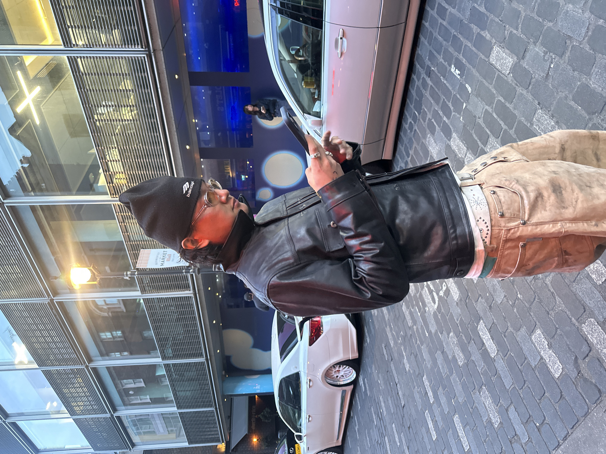 The image captures an urban street scene at what appears to be night or evening, indicated by the artificial lighting. The primary subject is a person, likely male, standing on a cobblestone street, engaged with a smartphone held in both hands. They are wearing a black beanie with "balenciaga" text and a logo, circular glasses, a black leather jacket, light brown or tan trousers, and a white studded belt over a light-colored shirt. Rings are visible on their fingers, and a tattoo can be seen on their left hand/wrist area.
To the left of the person is a modern building with a glass facade, reflecting ambient lights, internal structures, and a street lamp. Part of the text "BERWICK ST MARKET" is visible as a reflection on this glass surface, suggesting a location in London, United Kingdom. Behind the person, further down the street, is a white sedan car with custom white multi-spoke rims. Another individual, possibly female, is faintly visible in the background, reflected on the car's window and near a blue illuminated area with circular patterns on a building across the street. The ground is paved with grey cobblestones, some appearing lighter in color than others. The overall ambiance is that of a bustling city at night, with stylish elements and a focus on contemporary urban life.