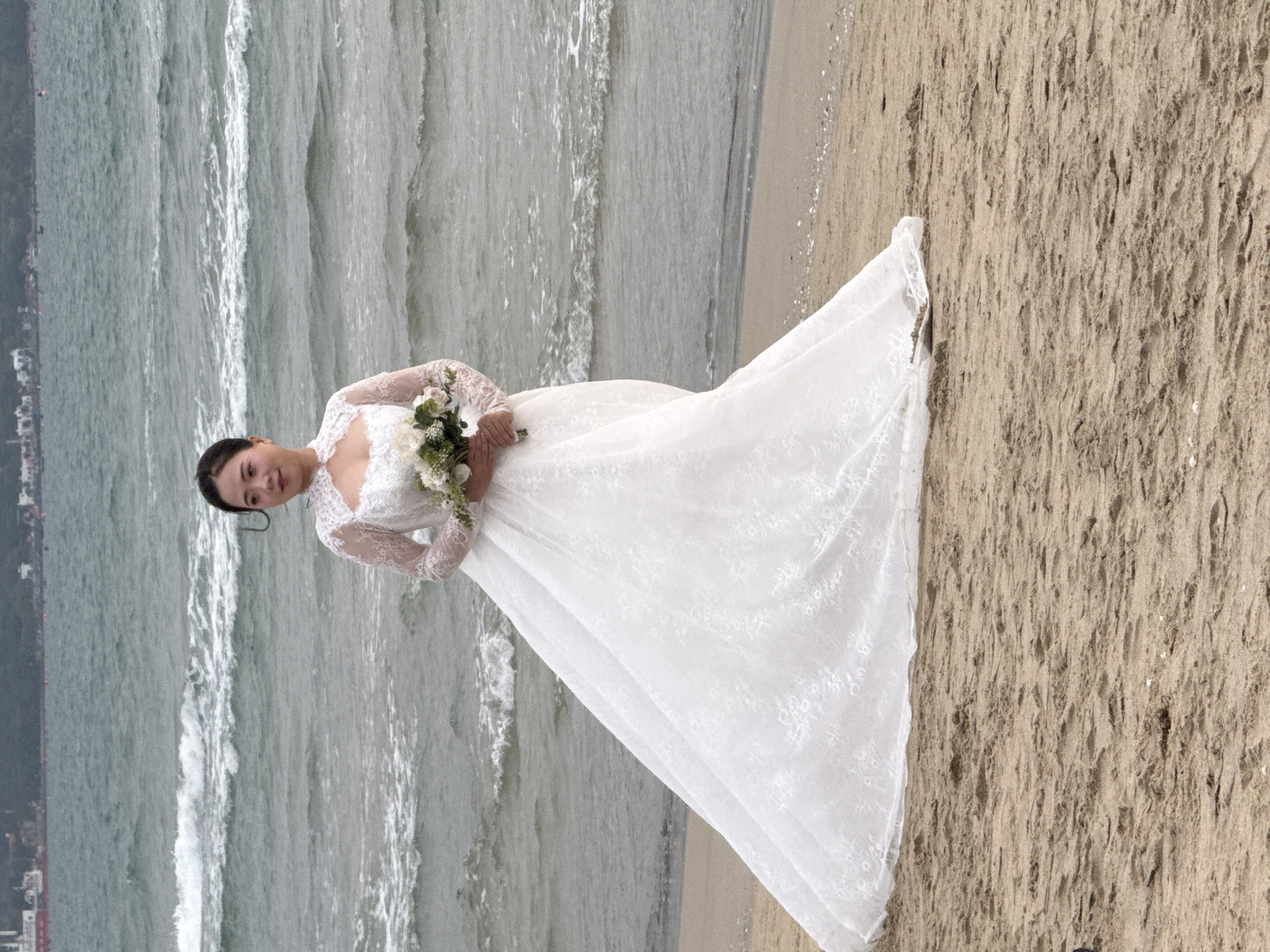 A woman in a white lace wedding gown stands on a sandy beach, holding a bouquet of flowers. The beach appears to be located in Đà Nẵng, Vietnam, based on the visible cityscape in the background. The ocean waves are visible on the left side of the image, creating a contrast between the water and the sand. The woman is facing the camera, and her gaze is direct, suggesting a formal portrait.