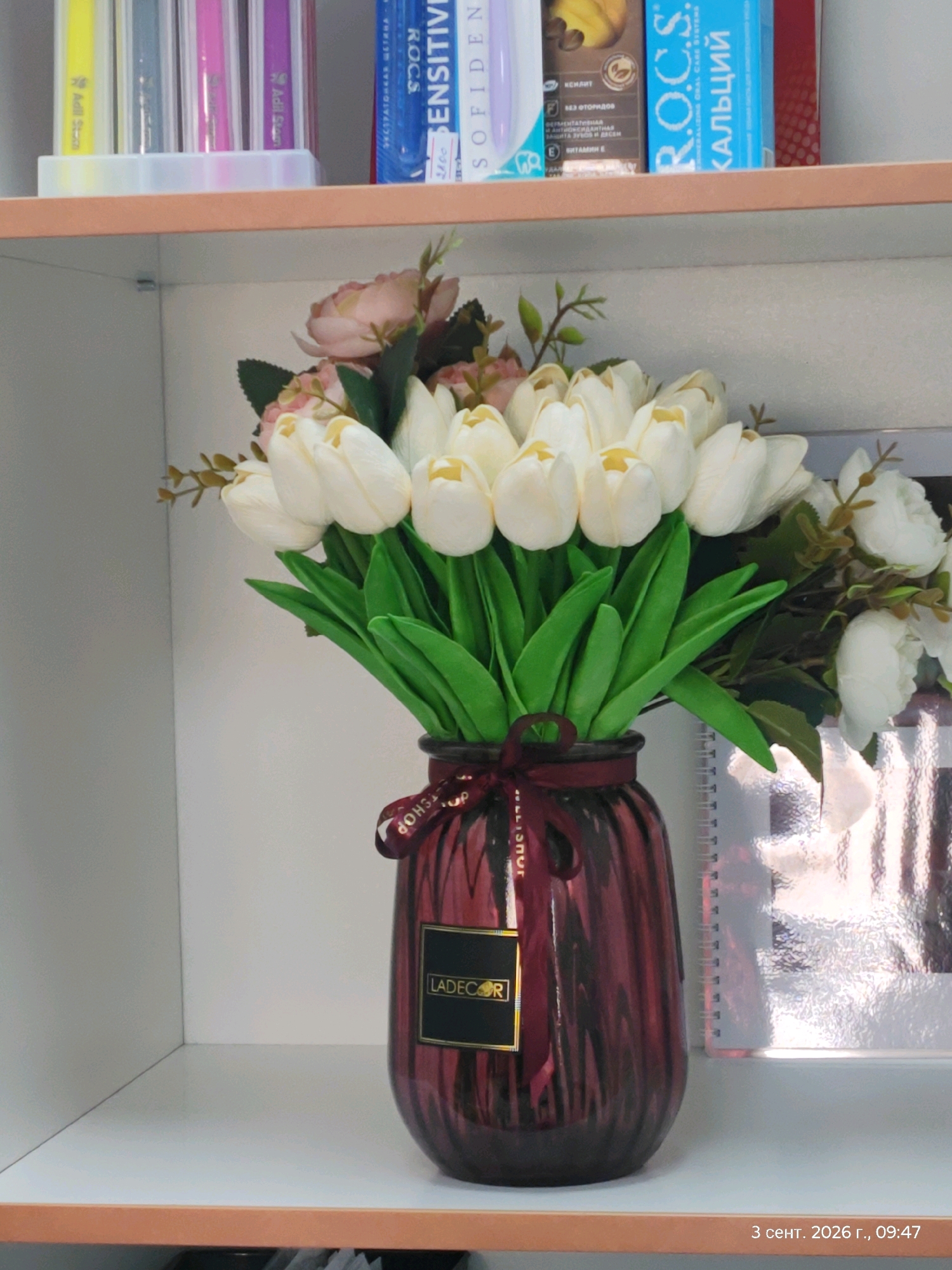 A close-up, eye-level shot captures a bouquet of artificial white tulips in a dark red ribbed glass vase. The vase is adorned with a dark red ribbon tied in a bow, featuring gold text. A black label with gold text that reads "LADECOR" is attached to the vase. Behind the tulips, several white artificial flowers and green leaves are visible. The bouquet rests on a white shelf, with an orange trim along its edge. To the left of the bouquet, partially visible on a higher shelf, are several colorful items, possibly pens or markers. To the right, a silver spiral-bound notebook with a textured cover is positioned next to the vase. The background is a plain, light-colored wall. The bottom right corner of the image displays a timestamp: "3 сент. 2026 г., 09:47". This scene appears to be an indoor setting, possibly a home or office environment, with no indication of the city of Astana, Kazakhstan.