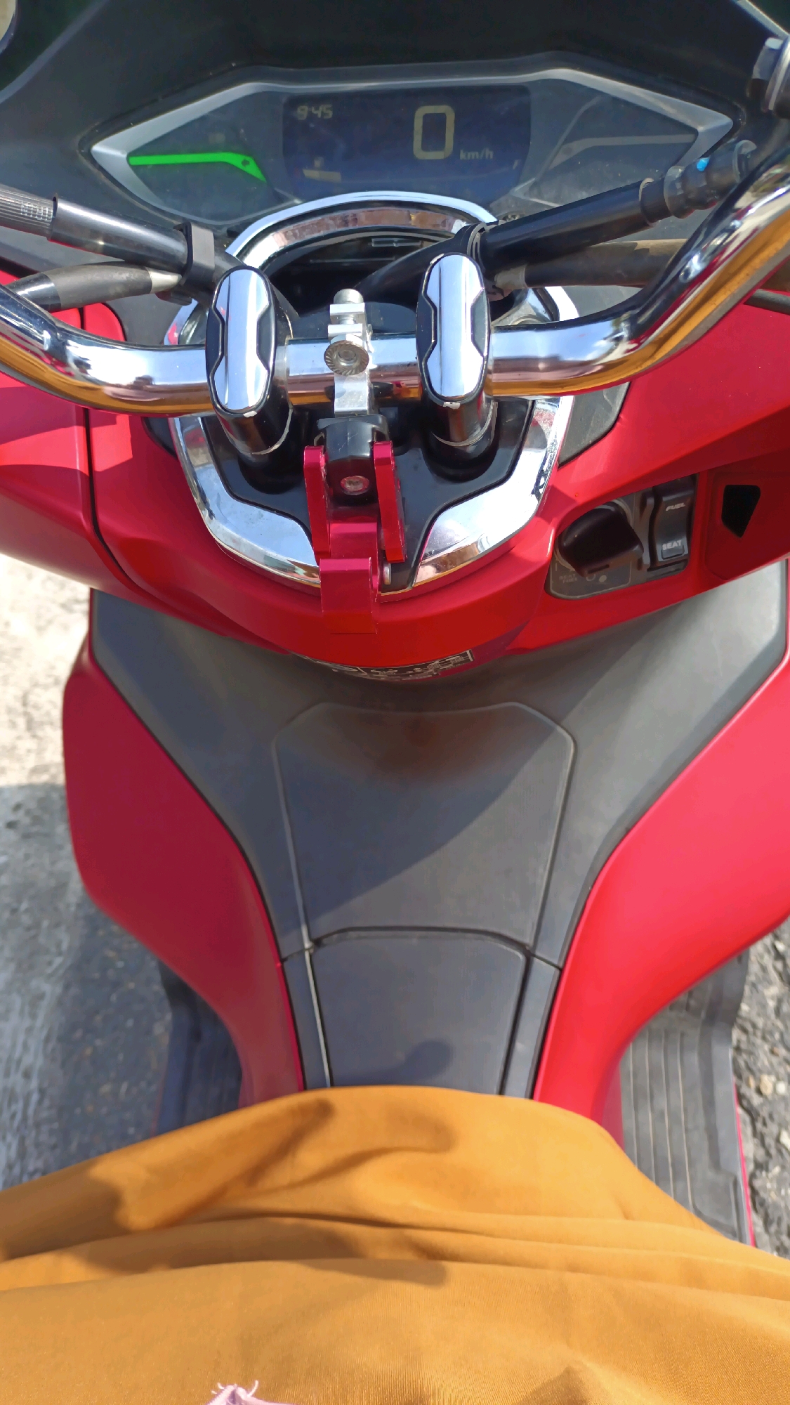 A close-up, overhead view of a red motorcycle is captured. The handlebars, dashboard, and front fairing are prominently displayed. The digital dashboard shows the time as 9:45 and the speed as 0 km/h. A chrome accessory is mounted on the handlebars. The motorcycle appears to be parked outdoors, with a textured ground surface visible. A portion of a person's leg, covered by an orange garment, is in the foreground, indicating the rider is seated on the motorcycle. The image is set in Panyalaian, Indonesia, suggesting a street or parking area within that city.