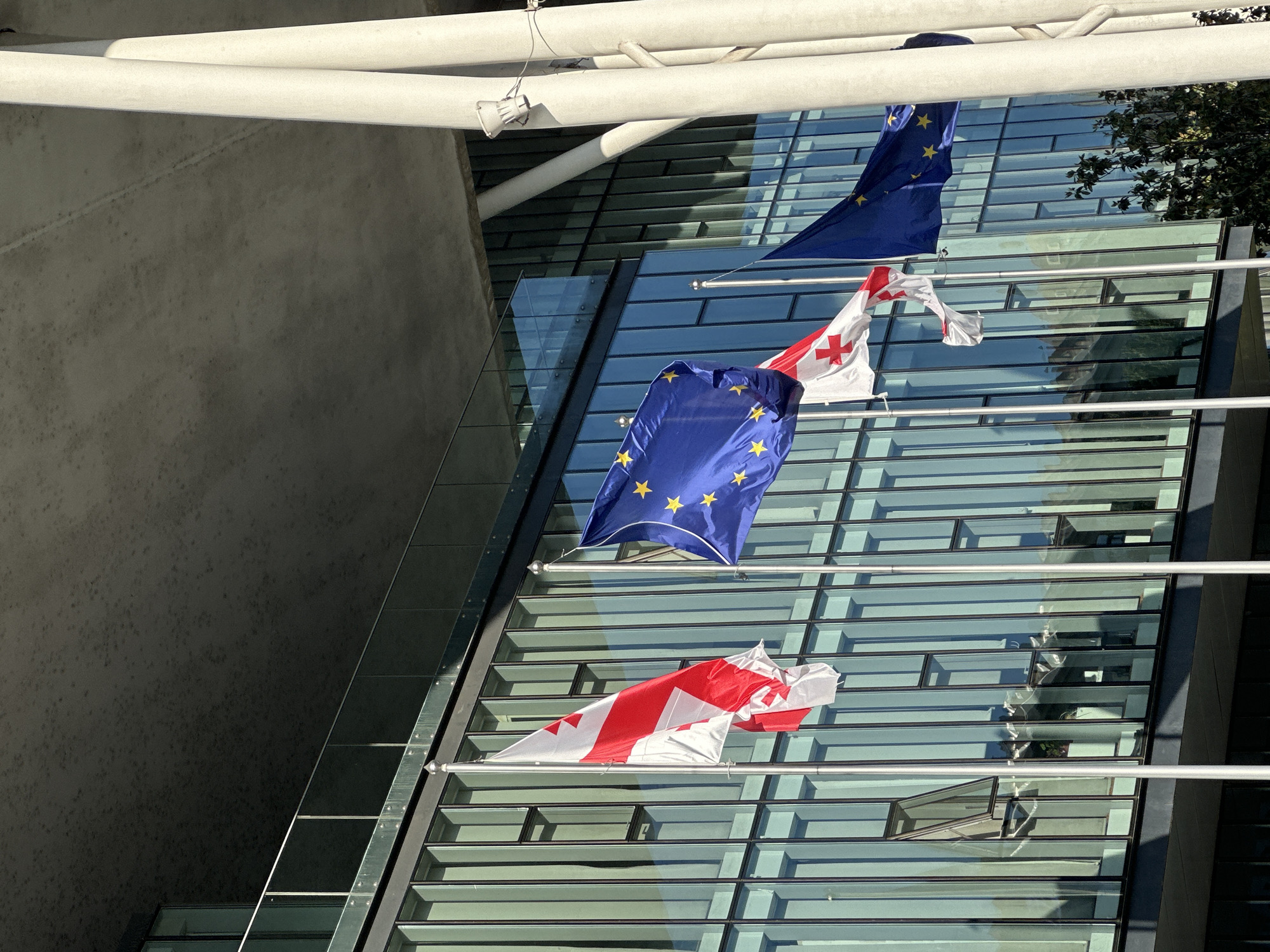 The image shows flags of the European Union and Georgia hanging from flagpoles mounted on the side of a modern building with glass windows. The flags are waving in the breeze. The building appears to be an office or government building. The location is unknown.