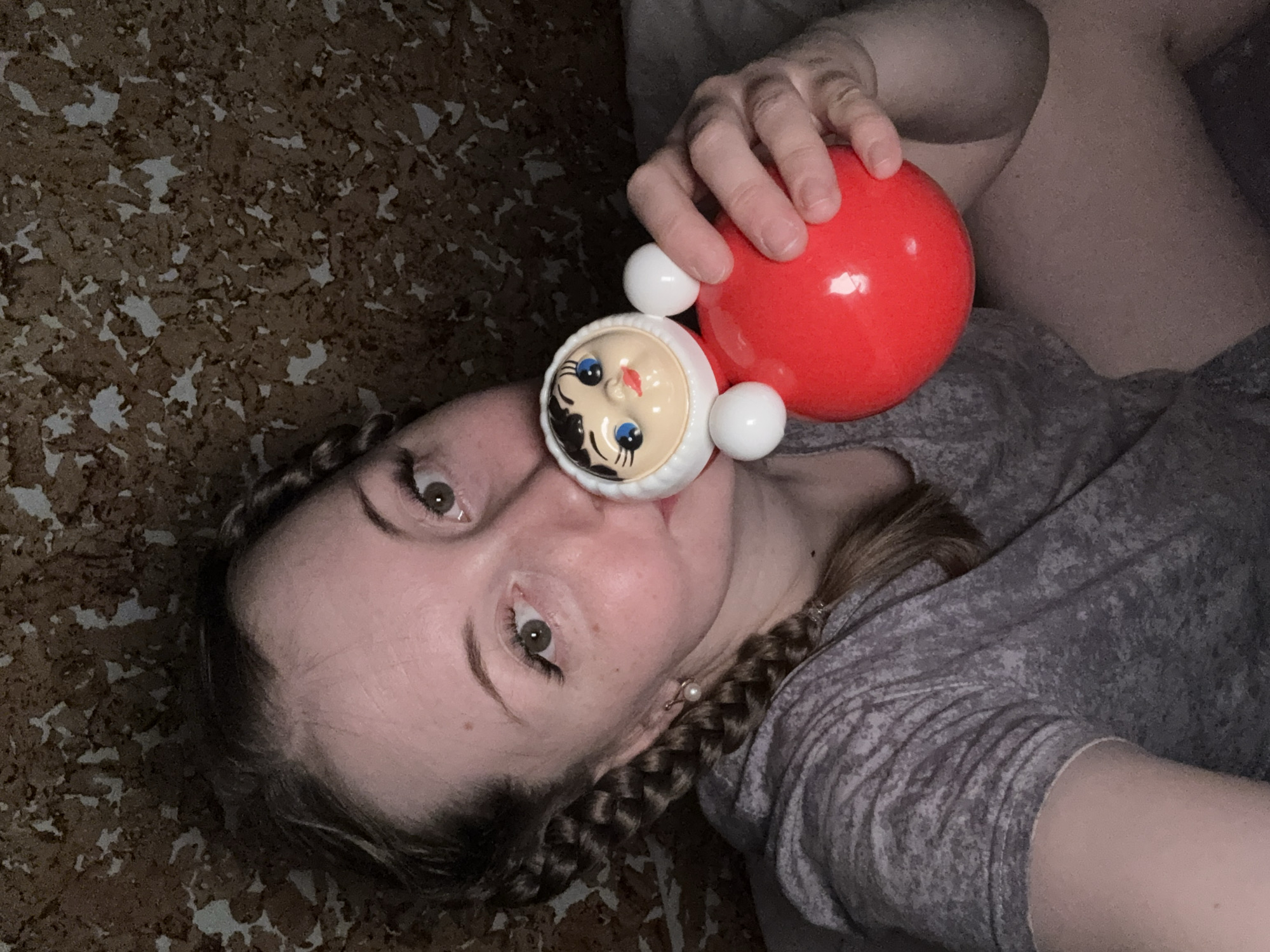 A medium close-up, high-angle selfie captures a woman lying down, interacting playfully with a roly-poly toy. Her head is tilted, and her light-colored eyes are open and directed towards the camera. The toy's white face, featuring blue eyes, red lips, and black hair details, is positioned close to or touching her nose and upper lip. She holds the toy's red spherical body with her right hand.
The woman has light-toned skin and light brown hair, which is styled in at least one visible braid resting on her left shoulder. She wears a grey t-shirt with a subtle pattern and a small, white pearl earring in her left earlobe.
The background is a textured surface with irregular brown and white/light grey patterns, resembling corkboard or a mottled fabric. The lighting is dim, suggesting an indoor setting with low light. There are no specific location cues beyond the patterned surface. Her expression is neutral to slightly playful. No text is visible in the image.