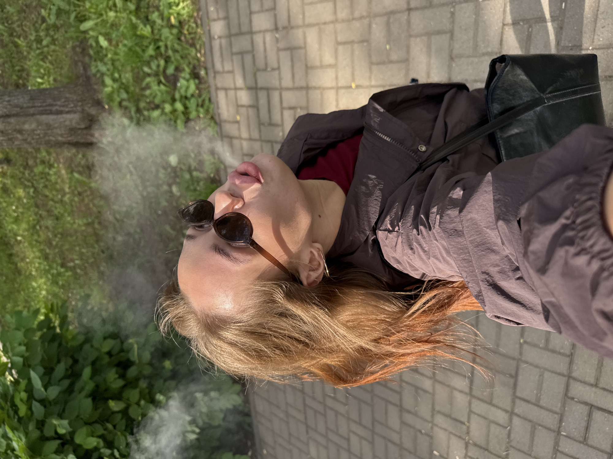 A young woman with light brown hair is captured in a selfie from a high angle outdoors. She is wearing sunglasses and a dark jacket over a maroon top. Smoke or vapor is visible near her face as she exhales upwards. The background shows a paved walkway, green foliage, and a tree trunk. The setting appears to be a park or a public outdoor area in Chaikovsky, Russia, as suggested by the overall ambiance and the time of day indicated by the lighting. A black handbag is slung over her shoulder.