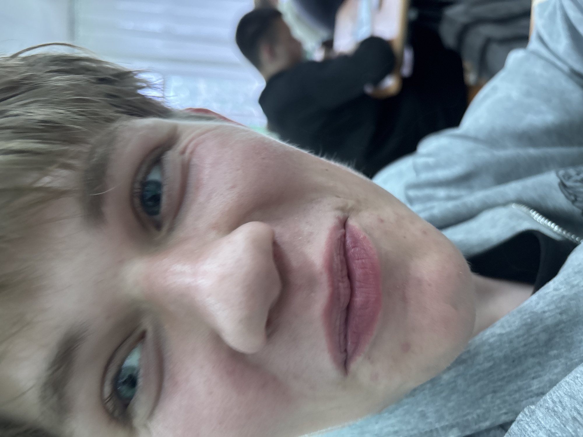 The image is a close-up, rotated selfie featuring a young person in the foreground within an indoor setting. The main subject, likely male, has light brown or blonde hair and blue eyes, and is looking directly at the camera with a neutral to slightly pouting expression. Their lips are faintly parted, and minor skin blemishes are visible on their face. They are wearing a grey hooded sweatshirt, with the zipper detail visible on the right side of the frame.
In the very blurry background, another individual, possibly male, can be seen seated at what appears to be a desk or table. This background figure wears a dark-colored top and is positioned with their back mostly to the camera. The overall setting suggests a casual indoor environment, such as a classroom or study area, given the presence of another person and the indistinct furniture. The lighting appears to be typical indoor illumination, offering no specific clues about the time of day or weather outside. There is no visible text in the image.