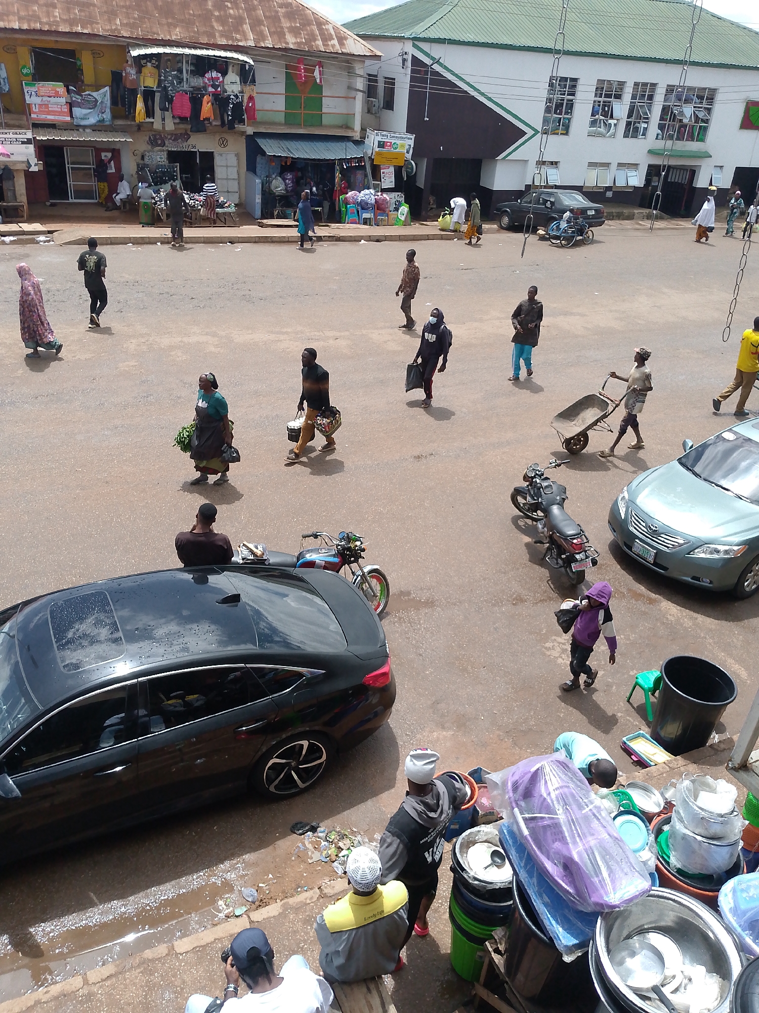 The image depicts a bustling street scene in Bukuru, Nigeria, likely a market area, with numerous individuals engaged in various activities. In the foreground, a black sedan is parked on the left side of the road, with its sunroof open. Next to it, several people are gathered, some appearing to be vendors selling wares such as pots and buckets. To the right of the sedan, a motorcycle is parked.
Further into the scene, a light green sedan is parked on the right. A man pushes a wheelbarrow across the paved road, indicating the movement of goods. Several other individuals are walking in different directions, some carrying bags or bundles. In the background, buildings line the street, including shops displaying clothing and other merchandise. The overall environment suggests a typical day in a Nigerian urban center, with a mix of commerce, transportation, and pedestrian activity. The weather appears to be overcast or hazy, with soft lighting.