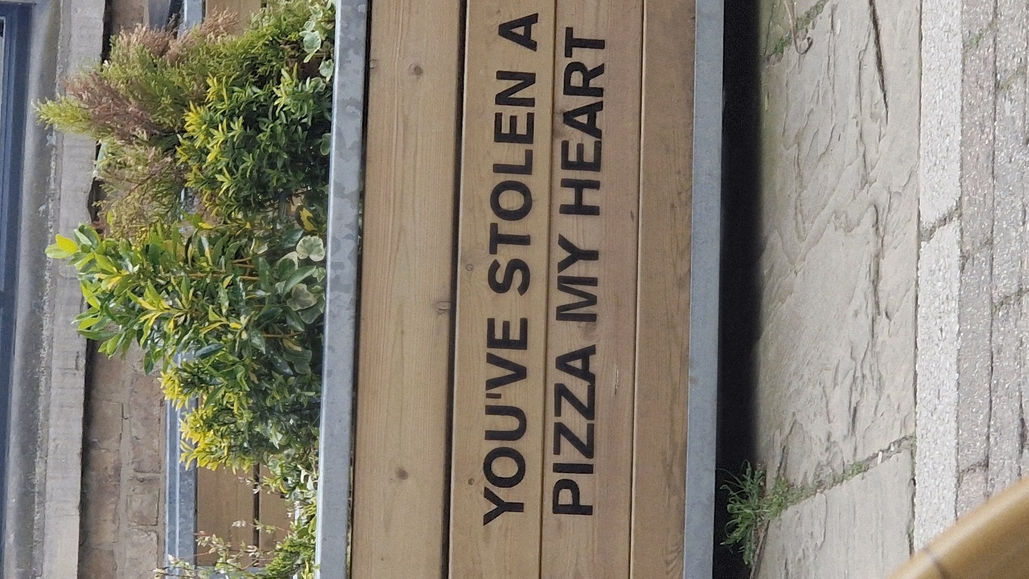 The image shows a vertical view of an outdoor setting. A rectangular wooden planter, filled with green plants, stands adjacent to a paved walkway. The planter has a metal frame. The words "YOU'VE STOLEN A PIZZA MY HEART" are printed in dark capital letters on the wooden side of the planter. The paving stones are light gray and textured. There is no visible evidence of human activity in the frame. The location can be inferred to be Lincoln, United Kingdom from the context.