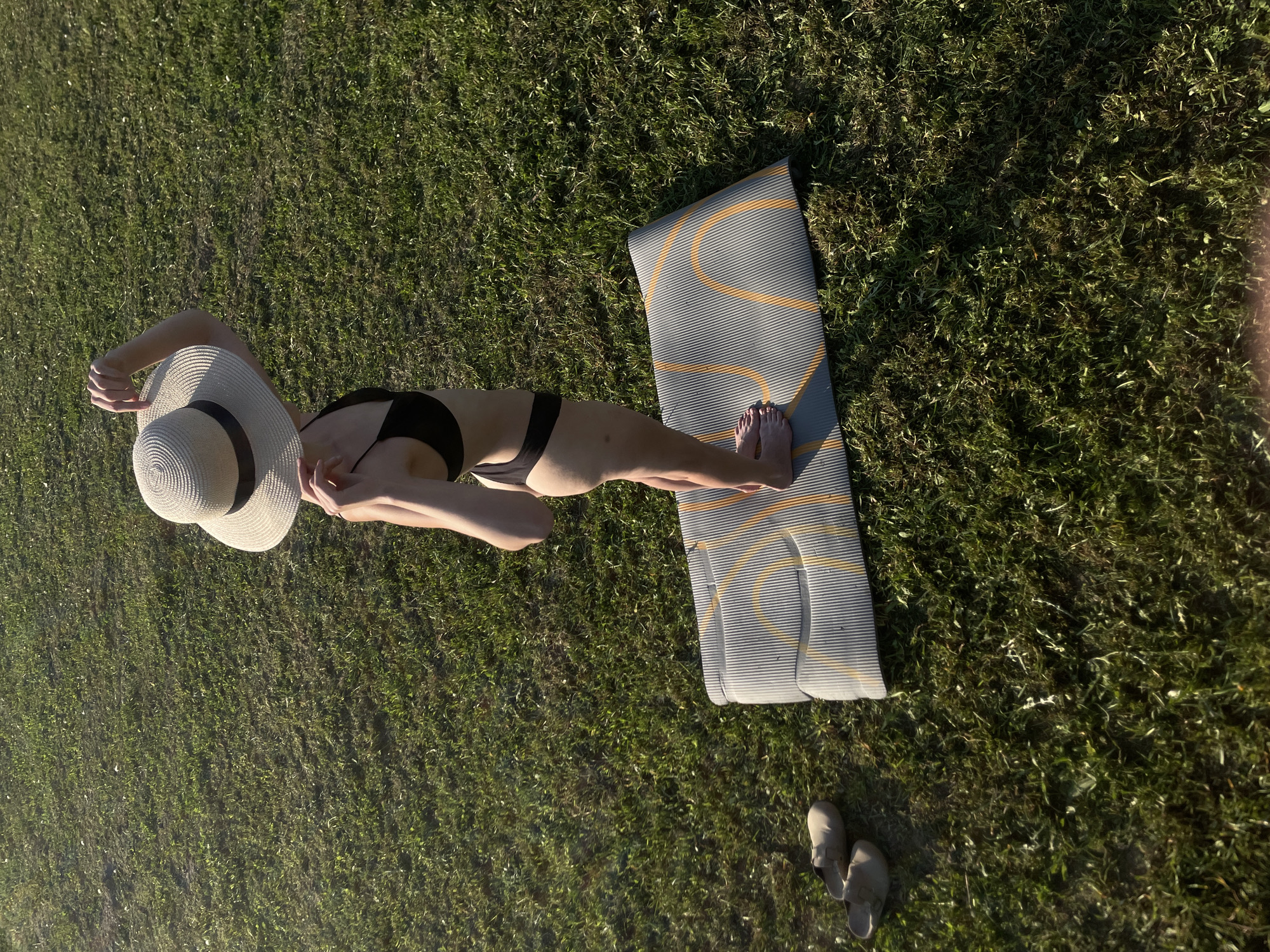 A high-angle shot depicts a woman in a black bikini and a wide-brimmed hat standing on a gray yoga mat with yellow stripes in a grassy area. Adjacent to the mat and woman are a pair of sandals. The scene appears to be in Подшивалово, Russia.