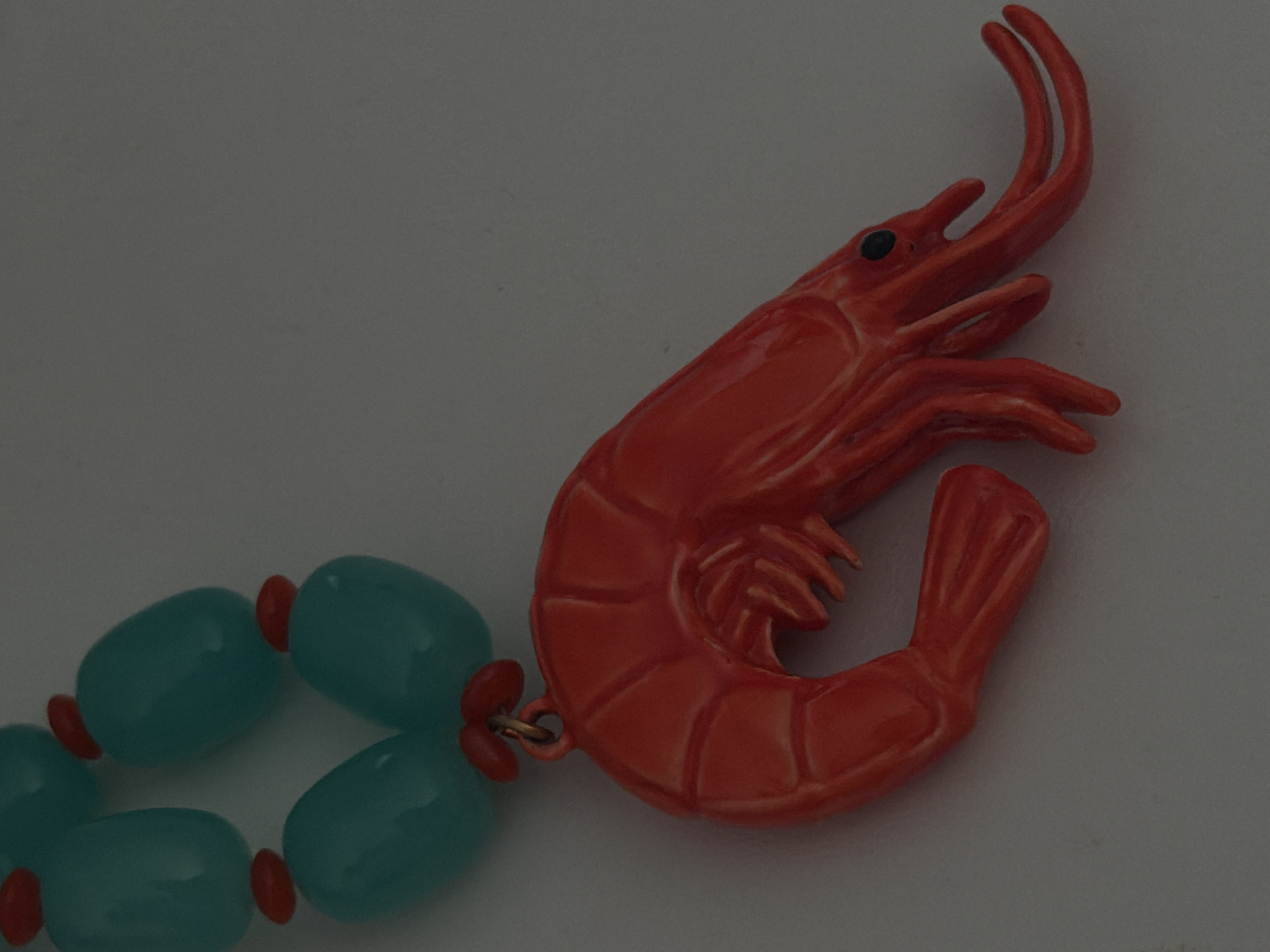 The image shows a close-up of a novelty necklace with a prominent orange shrimp pendant. The necklace is made of oval-shaped, turquoise beads interspersed with small, red, disc-shaped beads. The shrimp pendant is detailed, with segmented body segments, antennae, and legs, and appears to be made of a glossy material. The background is a plain, light-colored surface, possibly a table or countertop, with a slight shadow cast by the necklace. There is no indication of the city of P.im.sverdlova, Russia, or any people in the image.