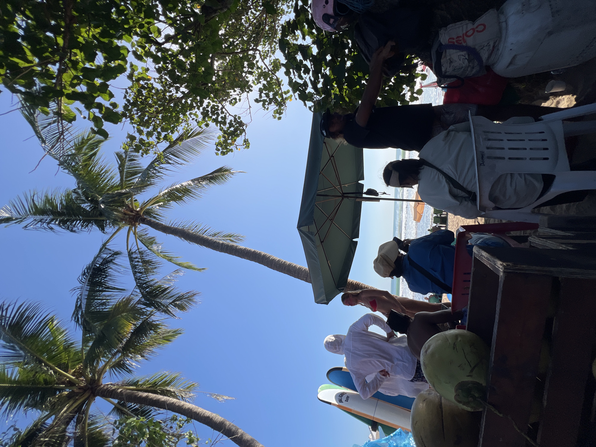 The image depicts a sunny day at Kuta Beach, Bali, Indonesia, near a beachside vendor. Two tall palm trees frame the clear blue sky. In the foreground, a wooden cart is filled with several green coconuts. To the left, a person in a white hooded jacket stands with their back to the camera, facing two surfboards lined up against the sand. Behind them, a woman in a red bikini is visible. Further to the right, a large green umbrella provides shade over a seated area where several people are gathered. One person in a dark shirt and baseball cap is interacting with another individual. Another person, seen from the back, wears a light-colored jacket and a headband, holding a phone. A white plastic chair is in front of this person. Towards the far right, a white cooler or storage container is present, with a bag on top. In the background, the ocean waves are gently rolling onto the shore, and a few beach chairs and umbrellas are set up further down the beach. The overall scene captures a relaxed atmosphere at a popular tourist destination.