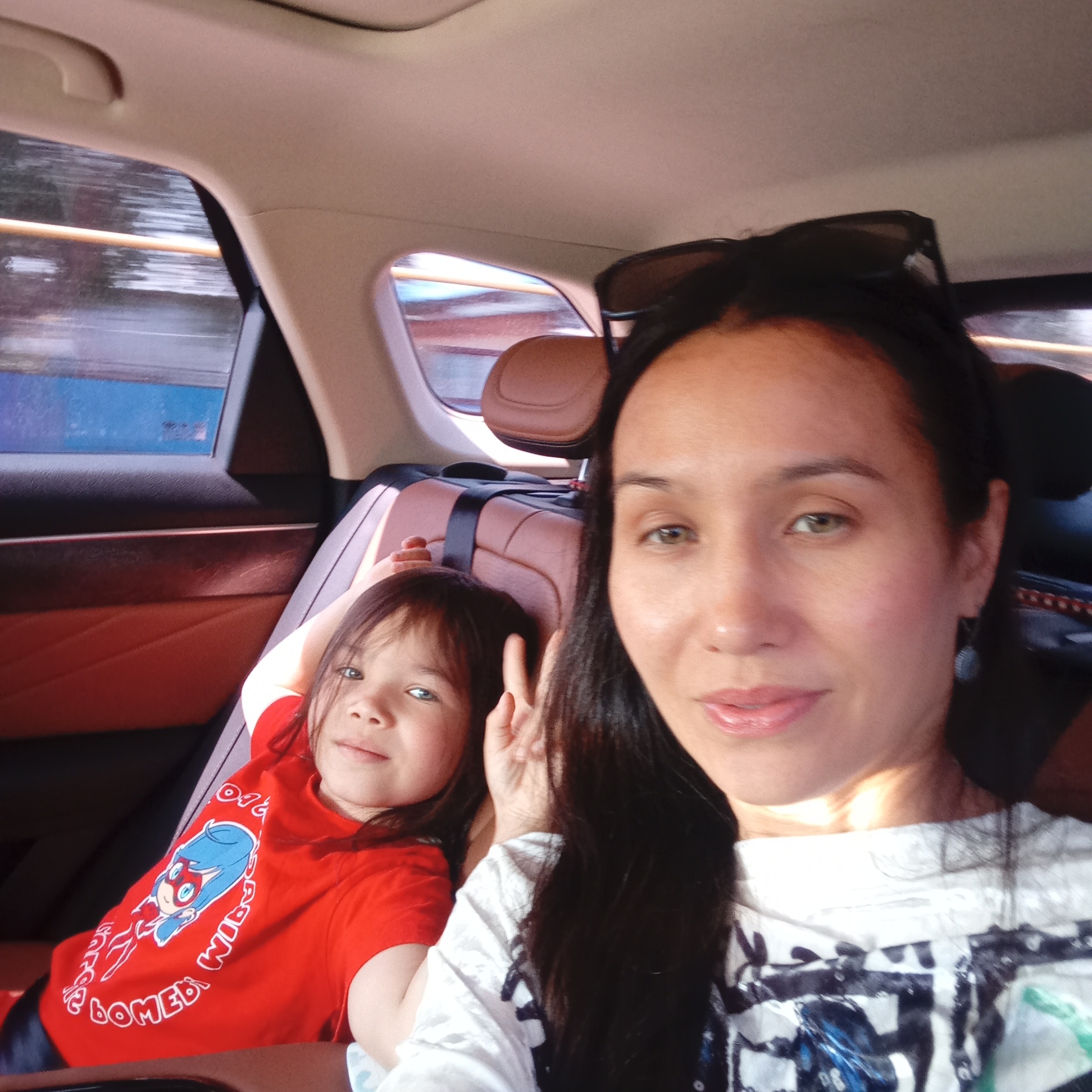 This is a selfie taken inside a car, likely in Izh-Boyki, Russia. A woman with dark hair and sunglasses perched on her head is in the foreground, looking towards the camera with a slight smile. She is wearing a white t-shirt with a black and blue graphic design. To her left and slightly behind her is a young child wearing a red t-shirt featuring a cartoon character. The child is also looking towards the camera and has their hand raised in a peace sign. The interior of the car is visible, with brown leather seats and the car's headliner. The window on the left shows a blurred outdoor scene of what appears to be a road with some structures, suggesting the car is in motion.