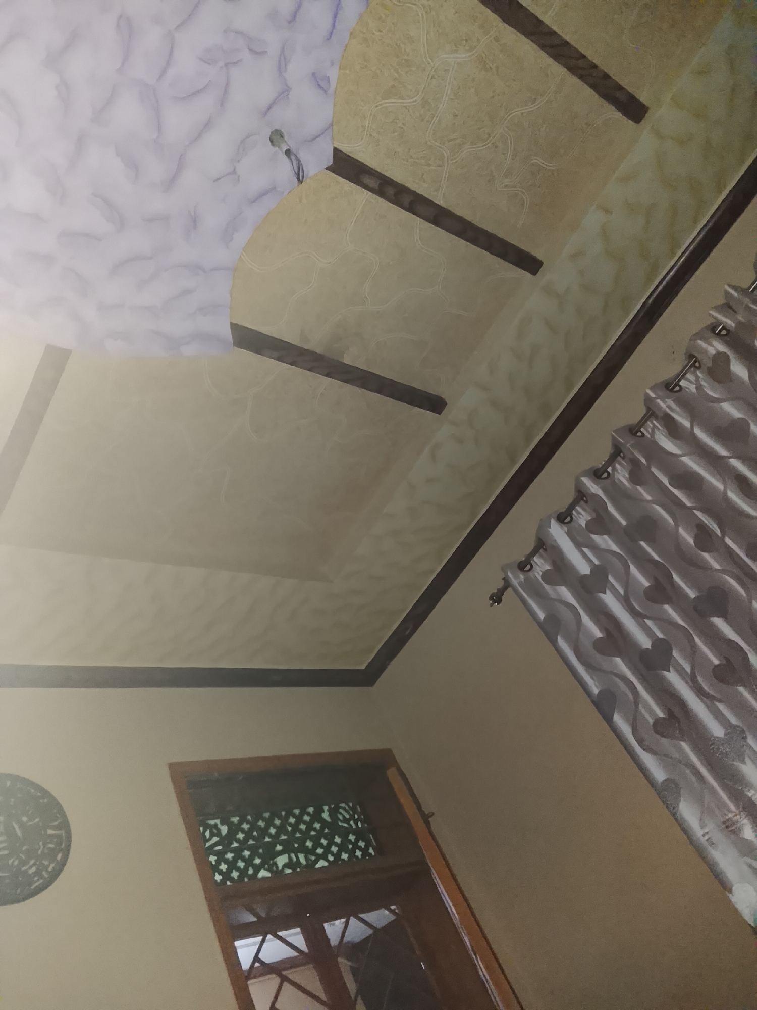 The image displays an indoor scene, likely a room or hallway, characterized by an elaborate false ceiling and neutral-toned walls. The ceiling features a multi-layered design with distinct sections. A prominent section in the upper left is light purple or lavender with a cloudy, mottled texture. Adjacent to it, a larger beige section is decorated with subtle, abstract, wavy line patterns. Dark brown or black trim pieces define these sections and create geometric patterns across the ceiling, including a central rectangular area of plain beige. A small, metallic light fixture or speaker is visible on the purple ceiling section.
To the right, light-colored curtains with a repeating heart pattern are hung on a metal rod, partially drawn. The walls are painted a uniform beige. In the lower left, a wooden door or window frame is visible, featuring an intricate lattice-work panel above the main opening. To the left of this frame, a circular, dark-colored wall hanging with detailed, possibly calligraphic or abstract, designs is mounted on the wall.
The lighting in the room is soft and even, suggesting either diffused natural light or interior artificial illumination. There are no people or activities taking place. The architectural and decorative elements, such as the false ceiling and lattice work, suggest a residential setting with possible South Asian or Middle Eastern design influences. No text is visible in the image.