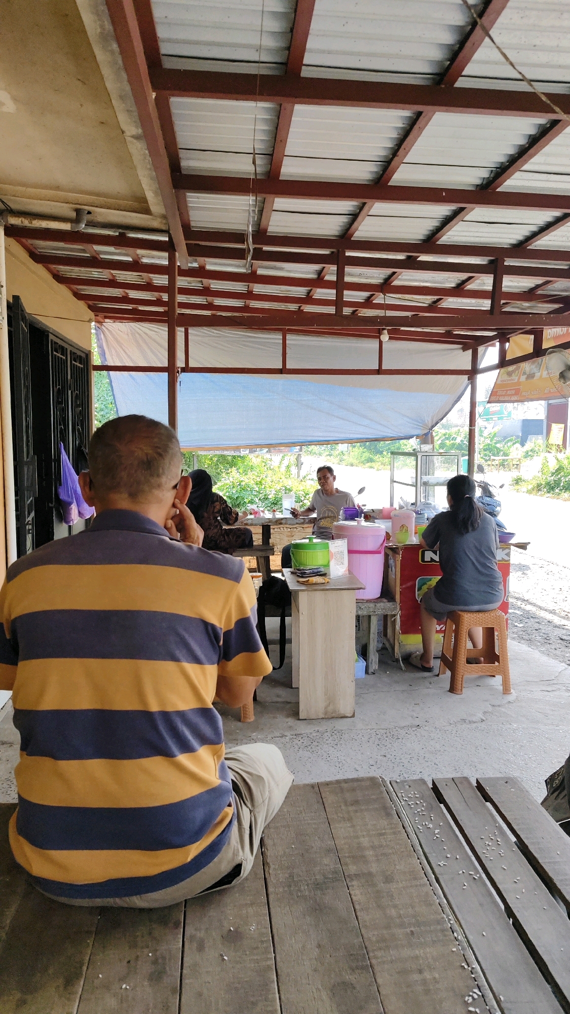 The image depicts an outdoor food stall in Banjarmasin, Indonesia, likely situated along a roadside given the presence of greenery and a road in the background. In the foreground, a man with a balding head, wearing a blue and yellow horizontally striped t-shirt, is seated facing away from the camera, with his hand to his ear. He is sitting on a wooden bench or platform constructed from planks.
Behind him, under a corrugated metal roof supported by a metal frame, several people are engaged in activities related to the food stall. A woman in a headscarf is seated on a bench, partially obscured. Another person, a man in a grey t-shirt, stands behind a stall counter, appearing to be serving or interacting with customers. He is holding a ladle and there are various containers and items on the counter, including a green cooler and a pink tub. To the right, a woman with long dark hair, wearing a dark grey t-shirt and shorts, is seated on a small orange stool at the counter. A motorcycle is visible parked behind the stall. The ground appears to be a mixture of concrete and dirt. The overall scene suggests a casual, open-air dining or take-away establishment.