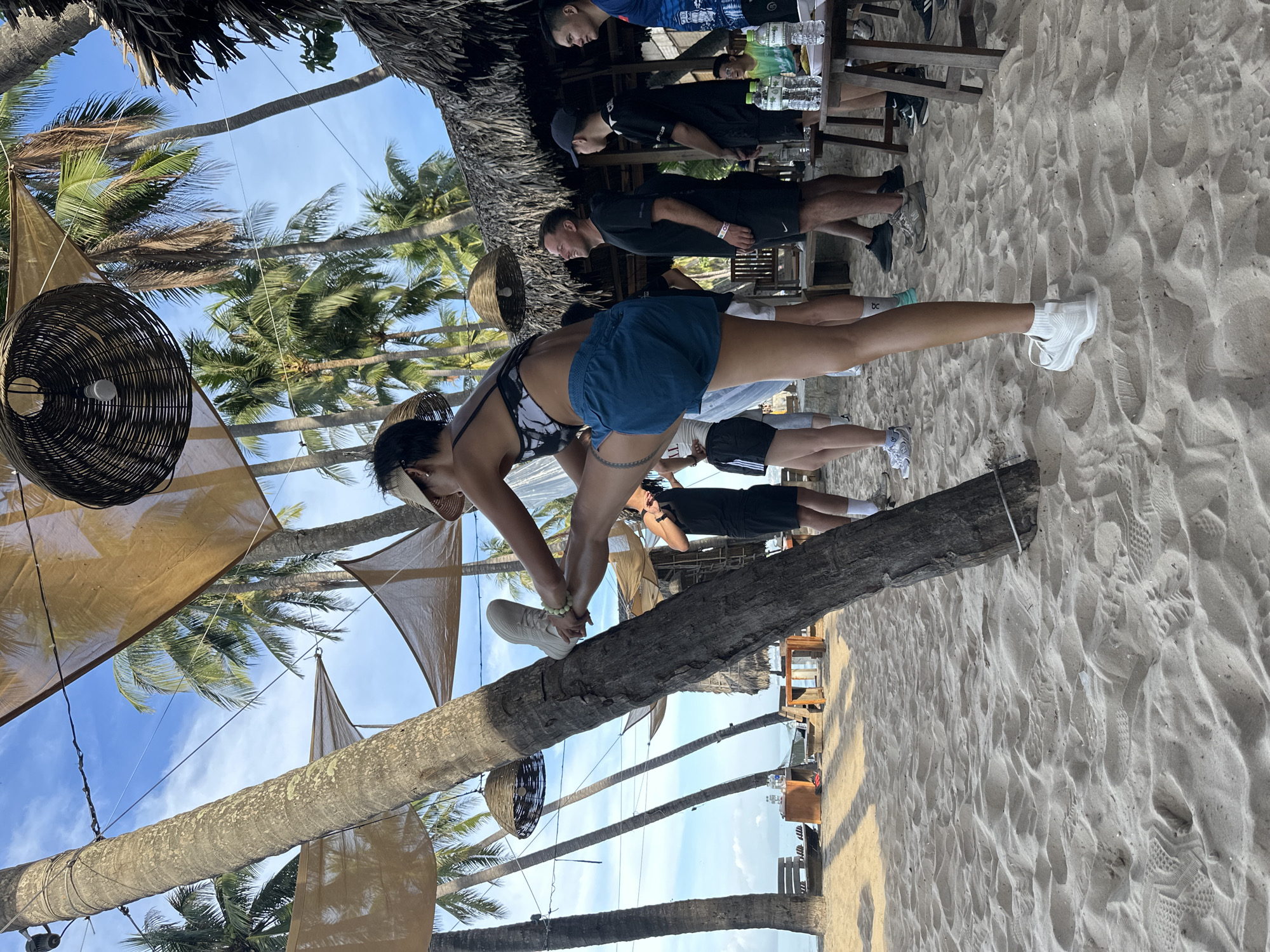 A woman is stretching her leg against a palm tree on a sandy beach in Mũi Né, Vietnam. She is wearing a black and white patterned top, blue shorts, and white sneakers. Her right leg is extended upwards and bent at the knee, with her foot resting on the trunk of the tree. She is wearing a visor and a bracelet.
In the background, several other people are standing or sitting on the beach. Some are wearing casual clothing, while others appear to be dressed for an activity. Palm trees surround the area, and triangular fabric shades are strung between them, providing shade. There are tables with bottles of water and other items on them. The scene suggests a relaxed outdoor gathering or event.