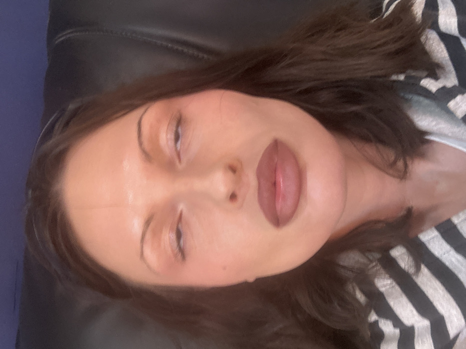 A female individual is centrally depicted in a close-up, chest-up portrait. She has dark brown hair, parted centrally, and her eyes are partially closed, with pupils obscured by the upper eyelids. Her lips are full and coated in a matte, muted reddish-brown cosmetic. She exhibits a subtle reddish flush on her cheeks. The subject is wearing a grey and white horizontally striped garment, visible on her upper body. The background consists of a dark, textured, upholstered surface, consistent with an interior setting. No other subjects or interactions are present. This scene is situated in Tunbridge Wells, United Kingdom.