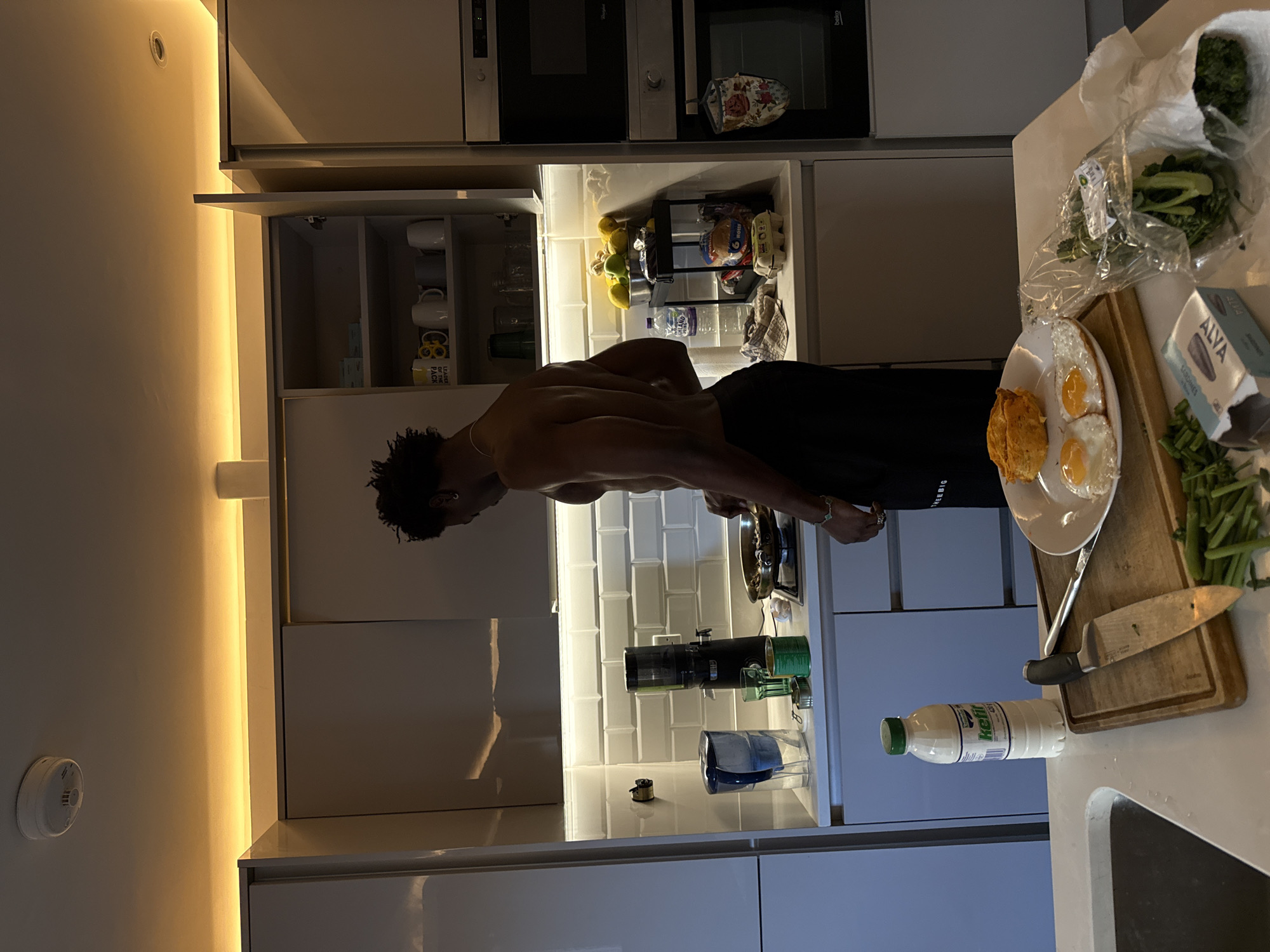 An individual with dark skin stands shirtless in a modern kitchen, wearing black shorts. They are positioned in front of a kitchen counter illuminated by under-cabinet lighting. The counter holds a plate with fried eggs and bread, a cutting board with a knife, green vegetables, a plastic bag of greens, and a carton of Alva sardines. A bottle of kefir is also visible near the edge of the counter. The kitchen has white cabinets, a built-in oven, and tiled backsplash. The location is within a residence, presumably in London, United Kingdom, based on the modern European kitchen design. The individual appears to be preparing or consuming a meal.