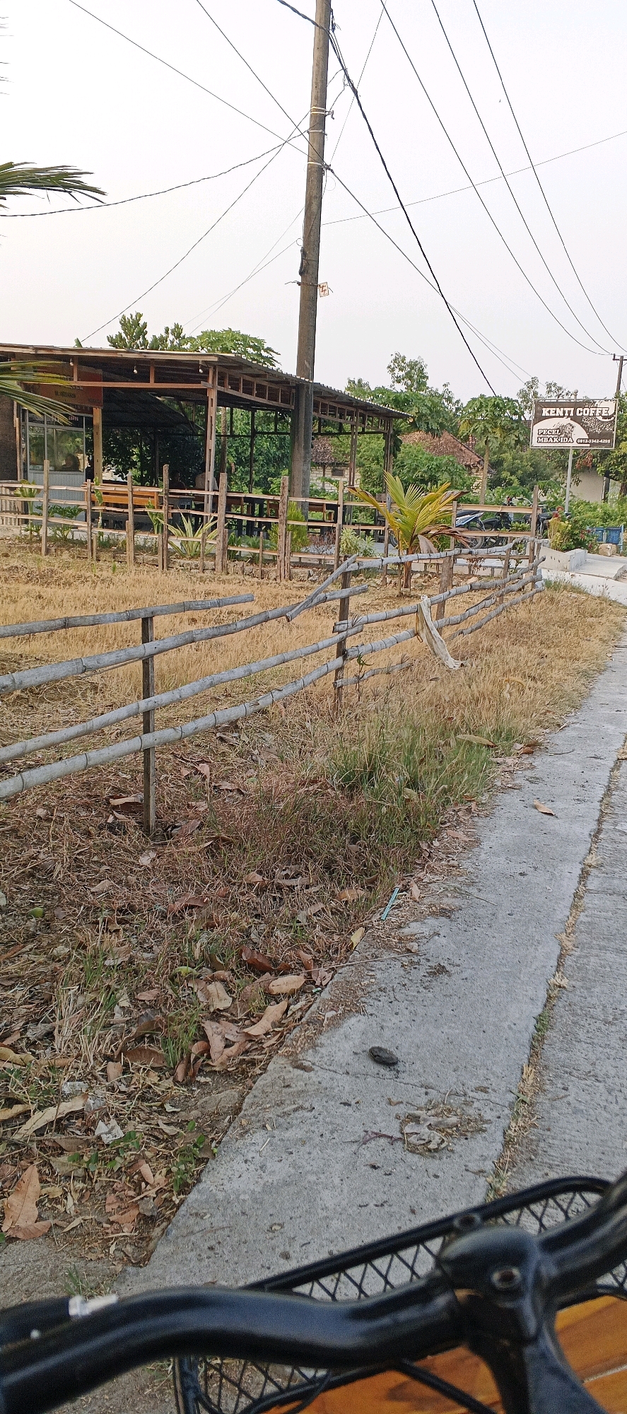 The image depicts an outdoor scene with a rustic, open-air establishment in Gumantuk, Indonesia. In the foreground, a portion of a bicycle's handlebars and basket is visible, suggesting the perspective of someone riding or stopped on a bicycle. A weathered bamboo fence runs horizontally across the middle ground, enclosing a dry, grassy field dotted with scattered leaves. Behind the fence, the establishment features a roof supported by wooden posts, with visible wooden tables and chairs. A signpost with text in what appears to be Indonesian, including "KENTI COFFE" and "PECEL MBAK IDA," is situated to the right, indicating a food and beverage venue. Electrical wires are strung across the sky, supported by a wooden utility pole. The overall environment is rural, characterized by dry vegetation and a simple, natural aesthetic.