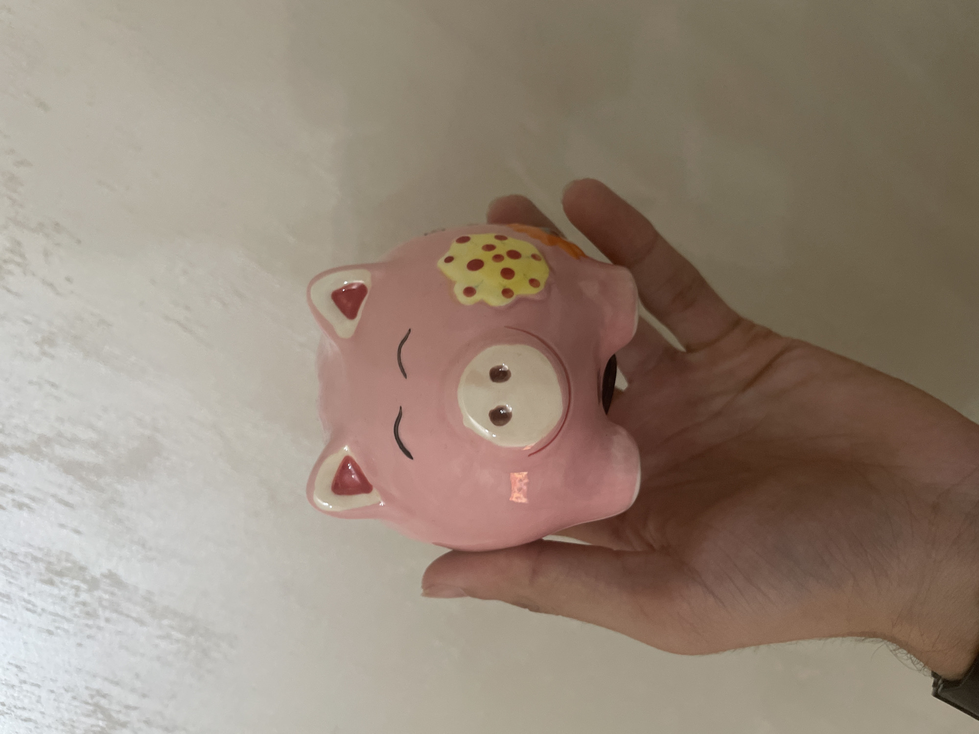 An upward-angled, close-up shot captured in the city of Ihtiman, Bulgaria, depicts a human hand holding a pink ceramic piggy bank. The piggy bank, centrally positioned, features a light pink glaze with distinct decorative elements. Its face displays two arched black lines for closed eyes, a raised white snout with two dark circular nostrils, and a subtle red smile line beneath the snout. The ears are pointed, with white outer edges and red inner triangles. On the piggy bank's left side (viewer's right), there is an irregularly shaped yellow patch adorned with multiple small red dots. A small, partial orange shape is visible on the right side of the piggy bank, near the base. The hand supporting the piggy bank is visible from the fingers curling around the base to the wrist, where a black watch strap is discernible. The background consists of a uniformly light, off-white or beige wall with a subtle, irregular texture, appearing out of focus.