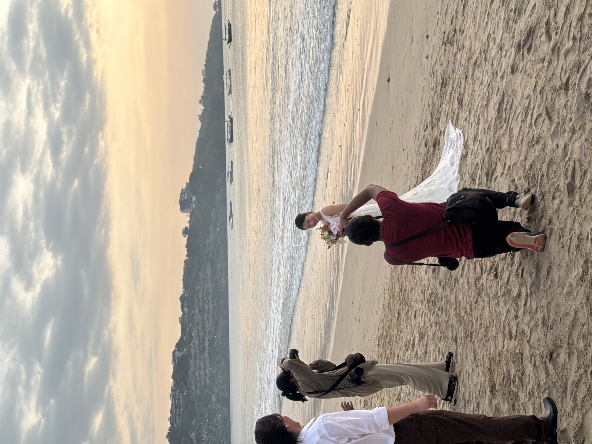 The image shows a wedding photoshoot on a beach in Kuta Selatan, Indonesia, at sunset. A bride, wearing a white wedding dress and holding a bouquet of flowers, is the central subject. Two photographers are actively taking pictures of her. One photographer is standing and holding a camera to their eye, while the other is kneeling, aiming their camera upwards at the bride. A fourth person, presumably part of the wedding party or staff, stands to the left, looking towards the bride. The beach has sandy terrain with some footprints and debris. In the background, gentle waves are lapping at the shore, and a line of boats are visible on the calm sea. The sky is filled with soft clouds and a gradient of orange and yellow hues from the setting sun, casting a warm glow over the scene. A lush, green hillside dotted with buildings and vegetation rises in the distance, characteristic of the landscape in Kuta Selatan.