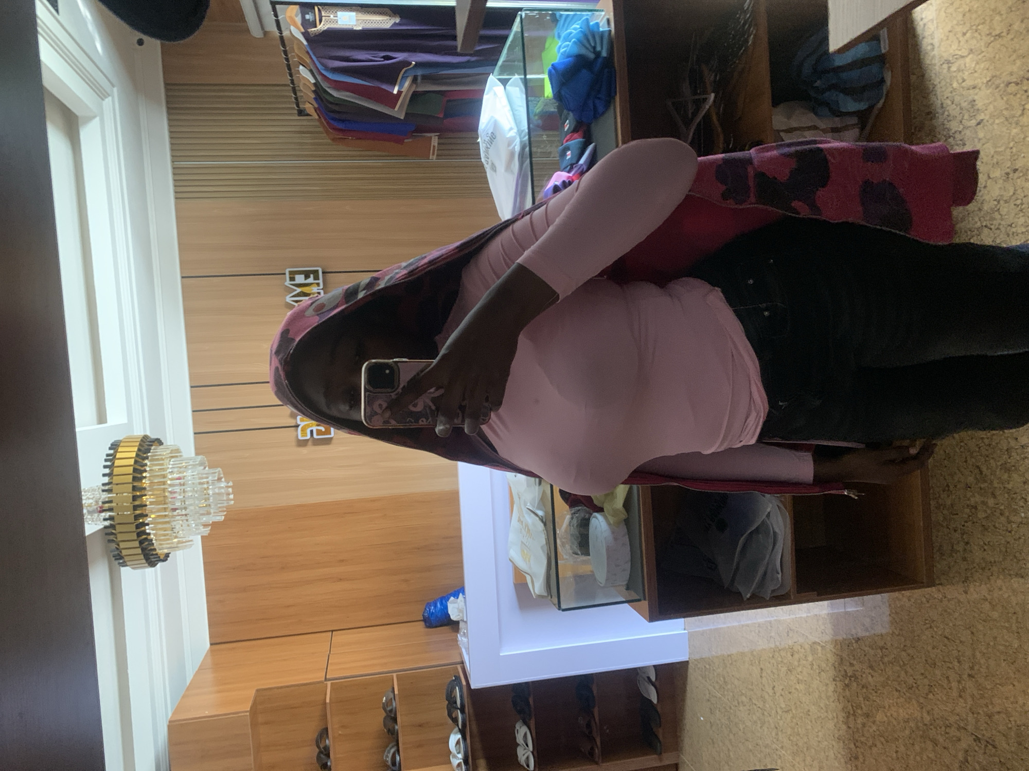 A person is taking a mirror selfie in a room that appears to be a retail space, possibly a clothing store. The person is wearing a pink long-sleeved shirt and dark pants. They are also wearing a head covering with a pink and dark patterned design. The room has wooden shelving units displaying items, a glass display case, and clothing racks with various garments. A chandelier is visible on the ceiling. The flooring appears to be a textured tile or linoleum. The environment suggests a commercial setting in Gura Top, Nigeria, likely a store selling clothing or related items.