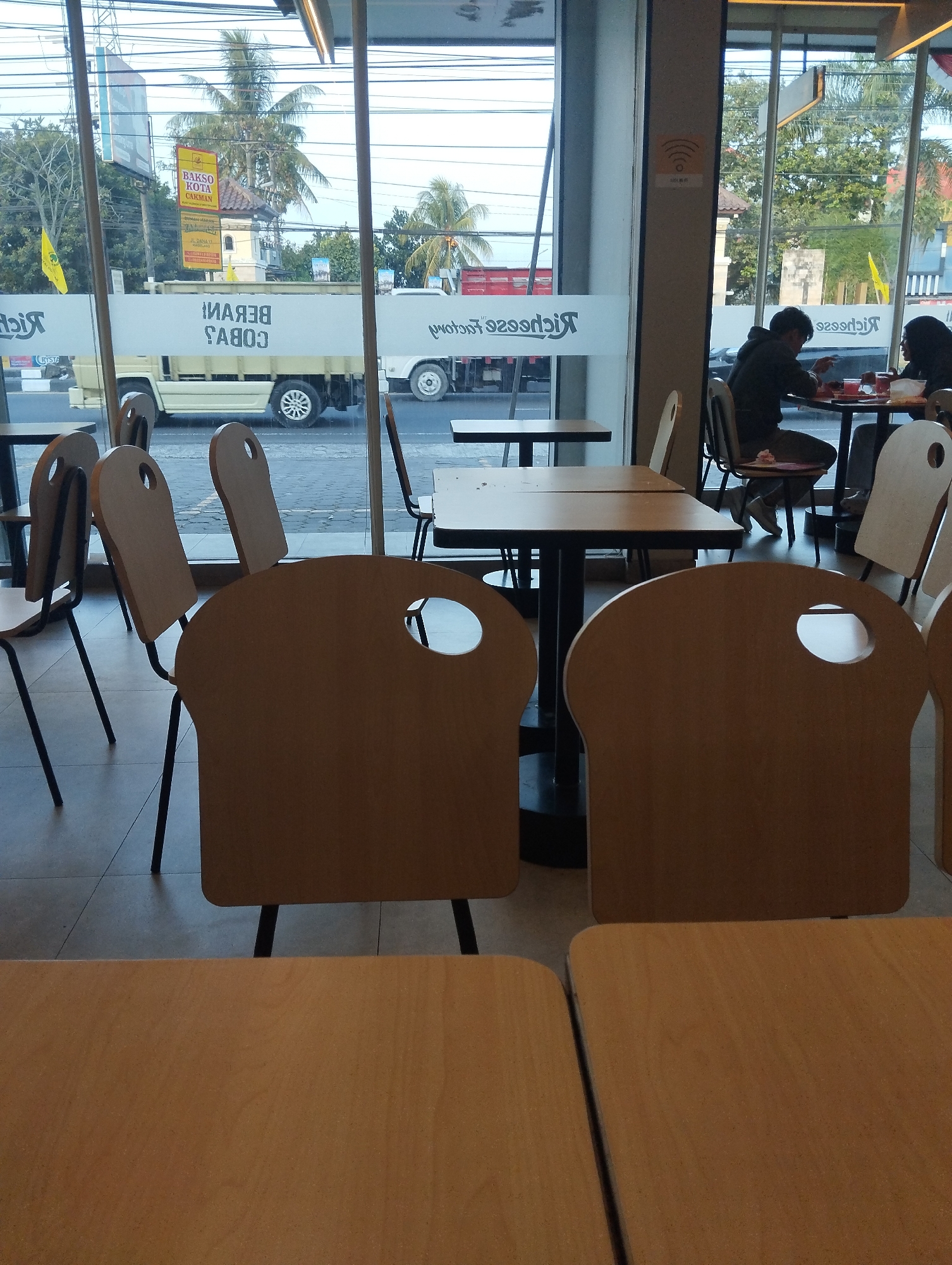 This image depicts the interior of a restaurant, likely a fast-food establishment, with large glass windows offering a view of a street in Sumberrejo, Indonesia. Outside, a light-colored delivery truck with the words "RICESSE COBA" visible on its side is parked on the road. Several palm trees and other greenery are also visible outside, along with a building displaying a sign that reads "BAKSO KOTA CAKMAN".
Inside the restaurant, several tables and chairs are arranged. Some chairs have a distinctive bread-loaf shape with a circular cutout in the backrest. Two individuals, a man and a woman, are seated at a table in the background, appearing to eat or interact. A Wi-Fi symbol is visible on a wall inside. The floor is tiled. In the foreground, the tops of tables and chairs are visible, suggesting the perspective is from a seated diner. The lighting suggests it is daytime.