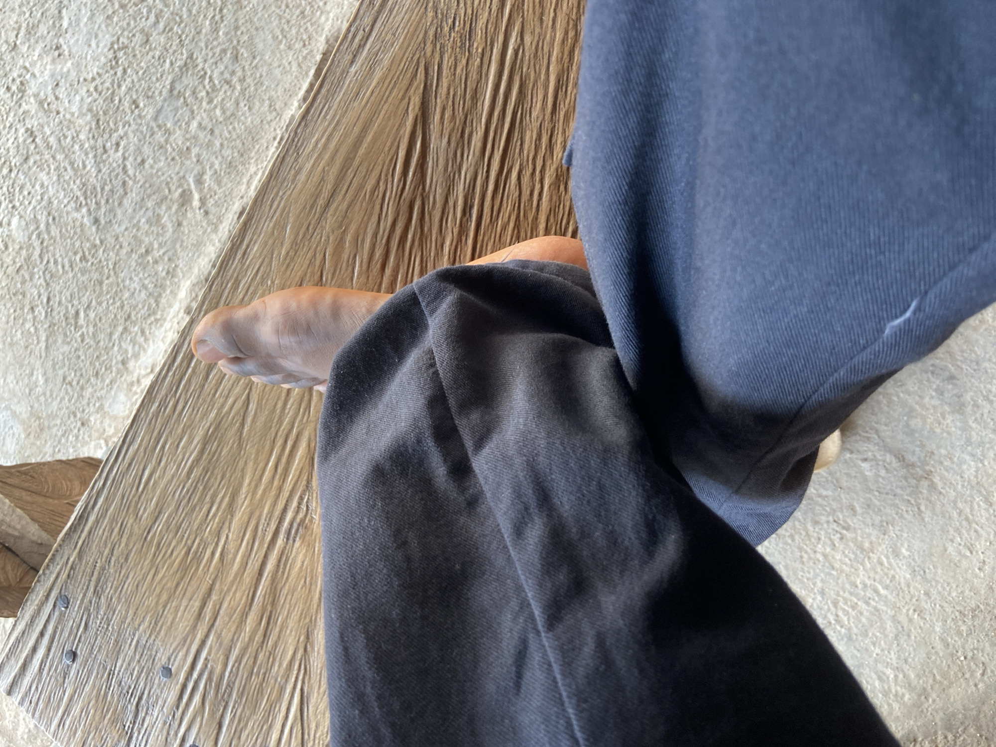 A high-angle, close-up shot captures a person's bare foot and lower leg, clad in dark blue or black trousers, resting in a casual setting. The person's right foot, visible from the toes to the ankle, has a medium-to-dark skin tone, suggesting a person of African descent, consistent with the given location context of Mararaba, Nigeria. The trousers are made of a durable fabric, possibly denim or canvas, and show significant creasing and folds.
The scene is dominated by two distinct background textures. To the left and upper right, there is a light-colored, rough, granular surface, which appears to be concrete, stucco, or a similarly textured floor or wall. Adjacent to this, and underneath the person's foot, is a prominent wooden surface. This appears to be a wooden plank or floorboard with a distinctive, deeply grooved, linear grain, giving it a somewhat rustic and worn appearance. Several dark, circular fasteners, likely nails, are visibly embedded in the wooden surface, particularly in the lower left portion of the wooden area, suggesting it is a fixed architectural element rather than a loose object. The two surfaces meet at a diagonal line across the frame.
The overall impression is one of a relaxed moment, with the person likely seated or reclining, allowing their foot to rest on or near the textured wooden surface. The bright and even lighting suggests it is daytime. There is no specific activity taking place other than resting. No visible text or discernible emotions are present in the image. The natural materials and simple textures align with a potential setting in Mararaba, Nigeria.