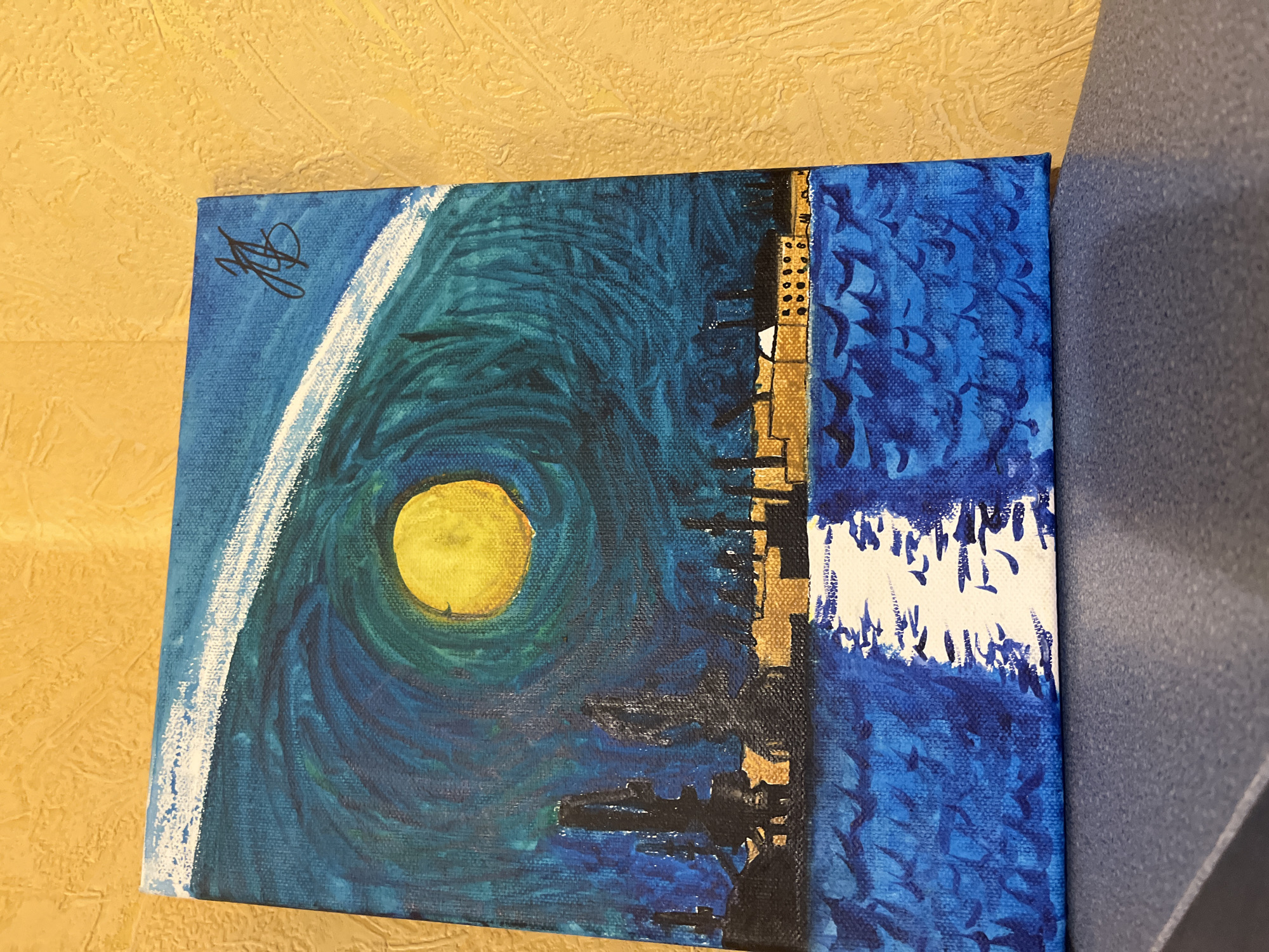 A rectangular canvas painting, oriented vertically, depicts a stylized nocturnal or crepuscular scene of a port city along a waterfront. The artwork is characterized by a strong color palette of deep blues, teals, and yellows, with elements rendered in black and white.
The upper two-thirds of the painting feature a swirling, turbulent sky in shades of deep blue and teal, creating a vortex-like effect. A prominent, large, bright yellow spherical celestial body, possibly a sun or moon, is positioned slightly to the left of the vertical center within the swirling sky, casting a strong luminescence. An arc of thick, textured white brushstrokes is visible in the upper right quadrant of the sky, extending downwards from the top edge. A stylized signature, composed of dark, connected initials "JB," is present in the upper right corner of the canvas.
Along the horizon, separating the sky and water, are silhouettes of industrial structures and buildings. On the left, several tall, dark, crane-like structures, indicative of a port, rise against the illuminated sky. To their right, a continuous line of low-lying buildings with rectangular forms and small, dark window-like details are depicted. These buildings feature illuminated yellow-gold facades or internal lighting, suggesting an active cityscape. This depiction of industrial port infrastructure and an urban waterfront is consistent with the characteristics of the Port of Burgas and the city of Burgas, Bulgaria.
The lower third of the painting portrays a body of water in various shades of blue, rendered with textured, choppy brushstrokes, suggesting waves. A distinct, elongated path of white light extends from the horizon line directly towards the viewer in the center of the water, reflecting the light from the celestial body above.
The canvas itself displays visible texture from the applied paint and the underlying fabric. The painting is resting upright on a dark blue-grey speckled surface, likely a shelf or table. The immediate background behind the painting is a uniformly textured yellow wall.