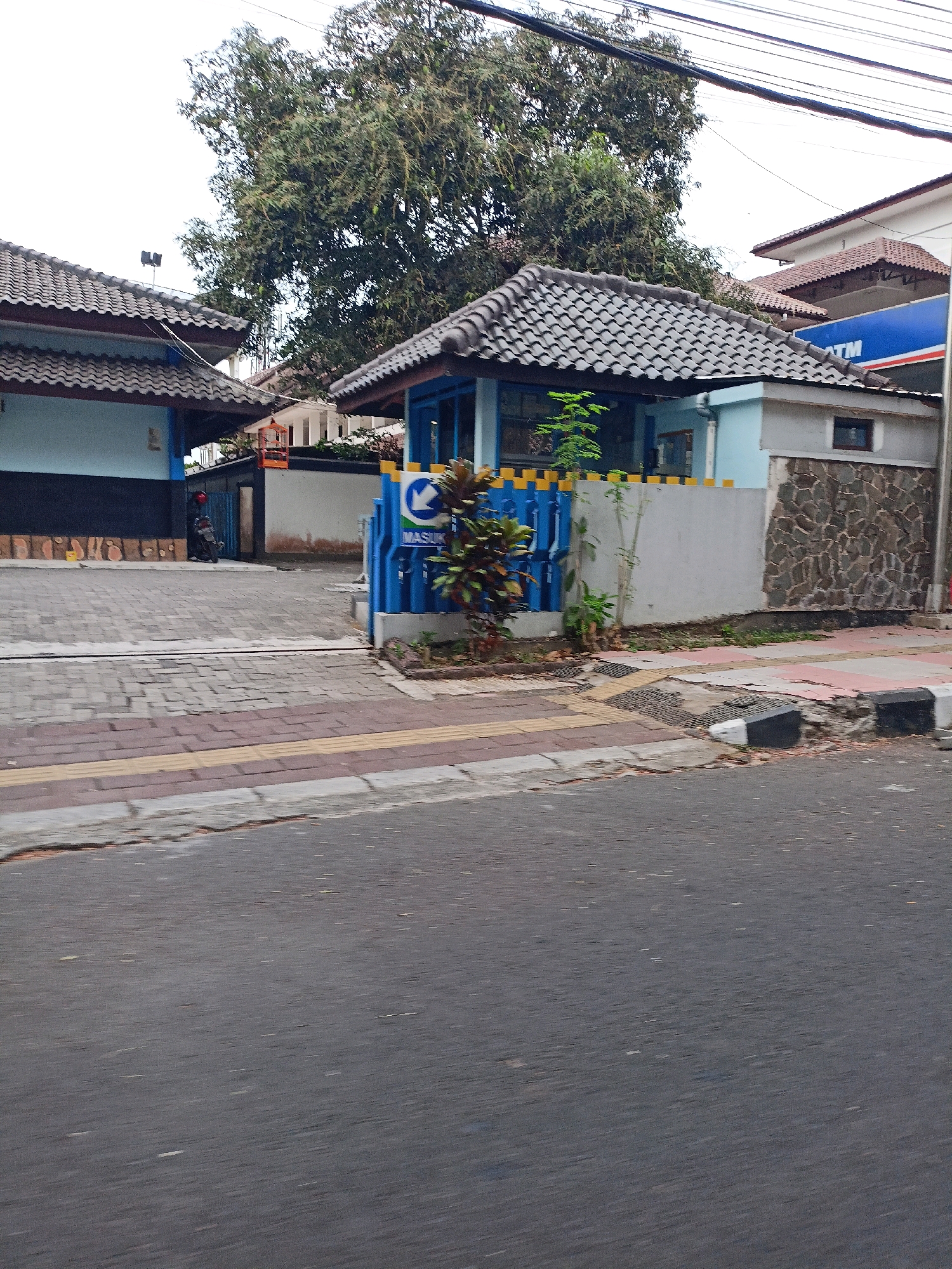 The image depicts the entrance to a property in Magelang, Indonesia, likely a security post or a small office. A blue and yellow striped gate is visible, with a sign displaying a white arrow pointing left and the word "MASUK" (Indonesian for "enter"). To the left of the gate, a building with a light blue facade and a dark tiled roof is partially visible. A motorcycle with a red helmet on the seat is parked near the entrance. Behind the gate, a grey wall with a stone texture forms part of the building. A large tree with dense foliage dominates the upper portion of the frame. The ground in front of the entrance is paved with bricks and has a yellow tactile paving strip. A street with asphalt pavement is in the foreground. The sky is overcast.