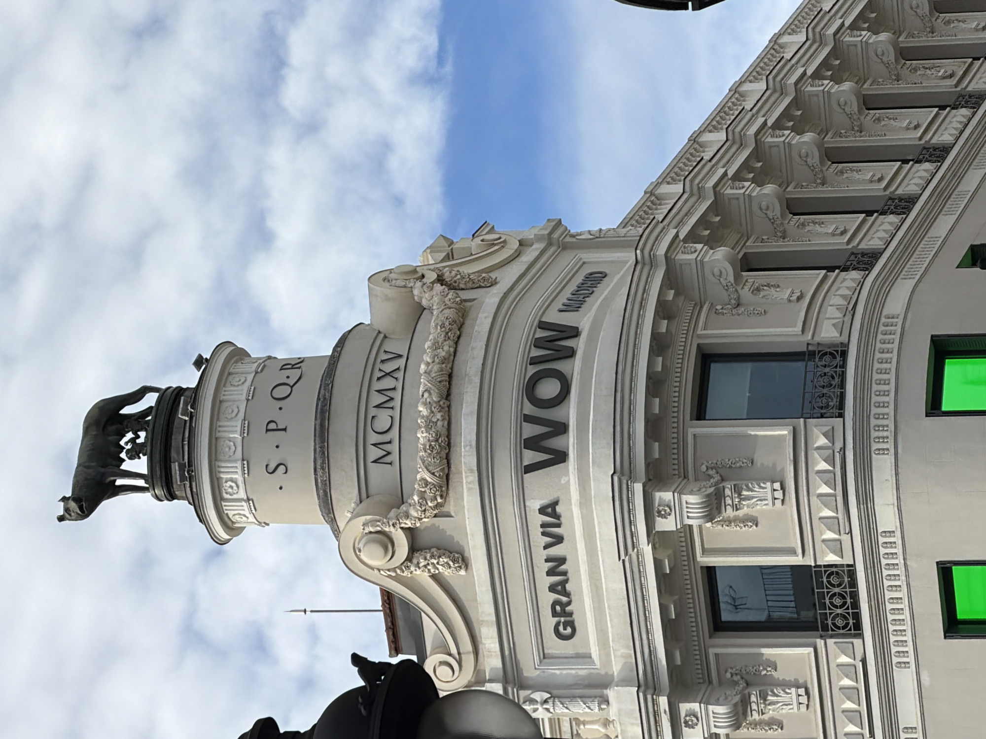 The image captures a portion of a building facade in Madrid, Spain. The building features ornate architectural details, including decorative moldings, window treatments, and sculpted elements. A prominent vertical structure extends upward from the building, adorned with the inscription "S.P.Q.R." and "MCMXV." At the top of the structure, a bronze sculpture depicts a she-wolf suckling Romulus and Remus, a symbol of Rome. The building displays the words "GRAN VIA" and "WOW MADRID" indicating the location or a business name. The sky in the background is partly cloudy.