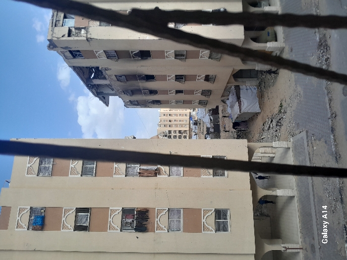 The image depicts a street scene in Khan Yunis, Palestinian Territory, viewed through vertical bars, possibly from a window or balcony.
In the foreground, a paved surface with gray interlocking pavers is visible. To the left, a multi-story, light-colored building stands mostly intact, featuring multiple floors with rectangular windows adorned by decorative arched frames. Laundry items, including blue fabric and darker clothes, hang from several windows on various levels. The ground floor of this building includes an arched colonnade where two individuals, dressed in dark clothing, are walking.
To the right, another multi-story, light-colored building of similar architectural style exhibits significant damage. Its upper floors show extensive structural collapse, with exposed rebar and a partially destroyed roof. Dark scorch marks and soot trails extend vertically down the facade from the upper levels, and numerous windows are broken or missing. The building's exterior shows widespread damage, including small impact points. A light-colored vehicle is partially visible under an arch on its ground floor.
Between these two primary buildings, the ground is unpaved and covered in rubble, dirt, and construction debris. Several makeshift structures, predominantly white tarpaulin tents, are erected in this area. Multiple small figures are visible within this debris-strewn space, with one individual in dark clothing walking near a prominent white tent, and other figures further in the background amidst the tents and rubble.
In the background, beyond the immediate damaged area, more multi-story buildings appear relatively intact under a sky with scattered white clouds against a clear blue. A "Galaxy A14" watermark is present in the lower-left corner of the image.