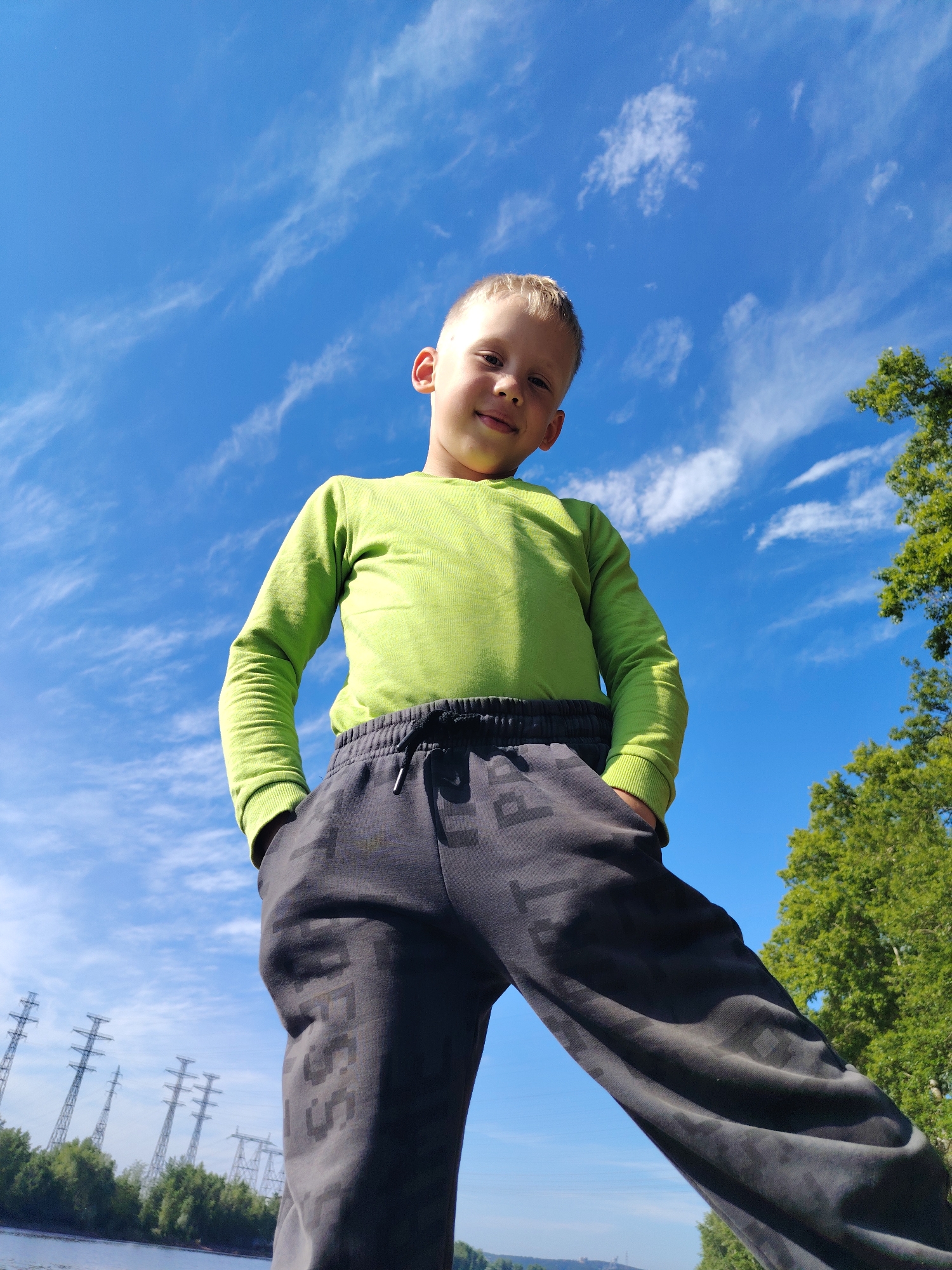 A low-angle outdoor shot captures a young boy, appearing to be around 6-10 years old, standing confidently and looking directly at the camera with a slight, pleasant smile. He has short blonde hair and is dressed in a long-sleeved, bright lime green shirt and dark grey sweatpants, with his hands casually tucked into his pockets. The sweatpants feature faint, dark grey lettering or patterns, with partial characters like "P" and "ESS" discernible.
The background is dominated by a clear, vibrant blue sky adorned with scattered, wispy white cirrus clouds, indicating a bright and sunny daytime setting. On the right side of the frame, the lush green foliage of a tree is visible, while across the bottom, a calm body of water, likely a river or lake, stretches horizontally. To the left, behind the water and distant trees, several tall, metallic electricity pylons (power line towers) rise against the sky, suggesting an area where natural landscapes meet infrastructure. This scene is consistent with an outdoor location near Chaikovsky, Russia, possibly along a significant waterway. The overall mood conveyed is cheerful and casual.