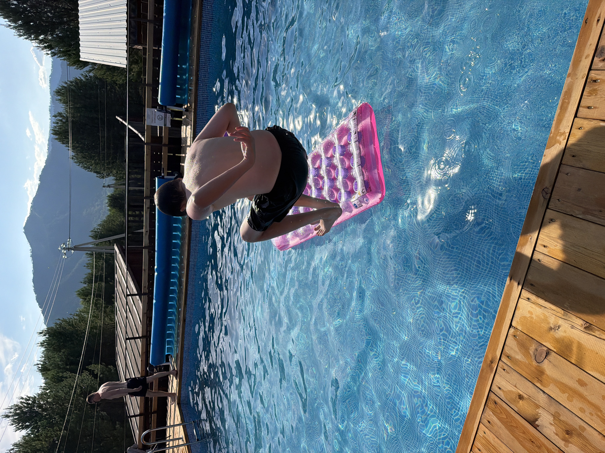 A young person is captured mid-air, about to enter a blue tiled swimming pool. They are wearing black swim trunks and their body is contorted as if they are jumping or diving. Beneath them is a partially inflated pink inflatable mattress with a hexagonal pattern. The pool water is clear and reflects sunlight, creating ripples and patterns on the surface.
To the left of the pool, there is a wooden deck with visible planks and screws. Beyond the deck, a grassy area leads to a wooden structure, possibly a fence or part of a building, and then a dense line of dark green trees. In the background, a mountain range rises, its slopes covered in trees and its peaks reaching towards a clear blue sky with some wispy white clouds. The lighting suggests it is daytime, likely sunny. A small white sign with black text is attached to the wooden structure. The overall scene depicts a recreational activity in an outdoor setting, likely a warm day given the swimming pool activity. The location context provided is Elekmonar, Russia.