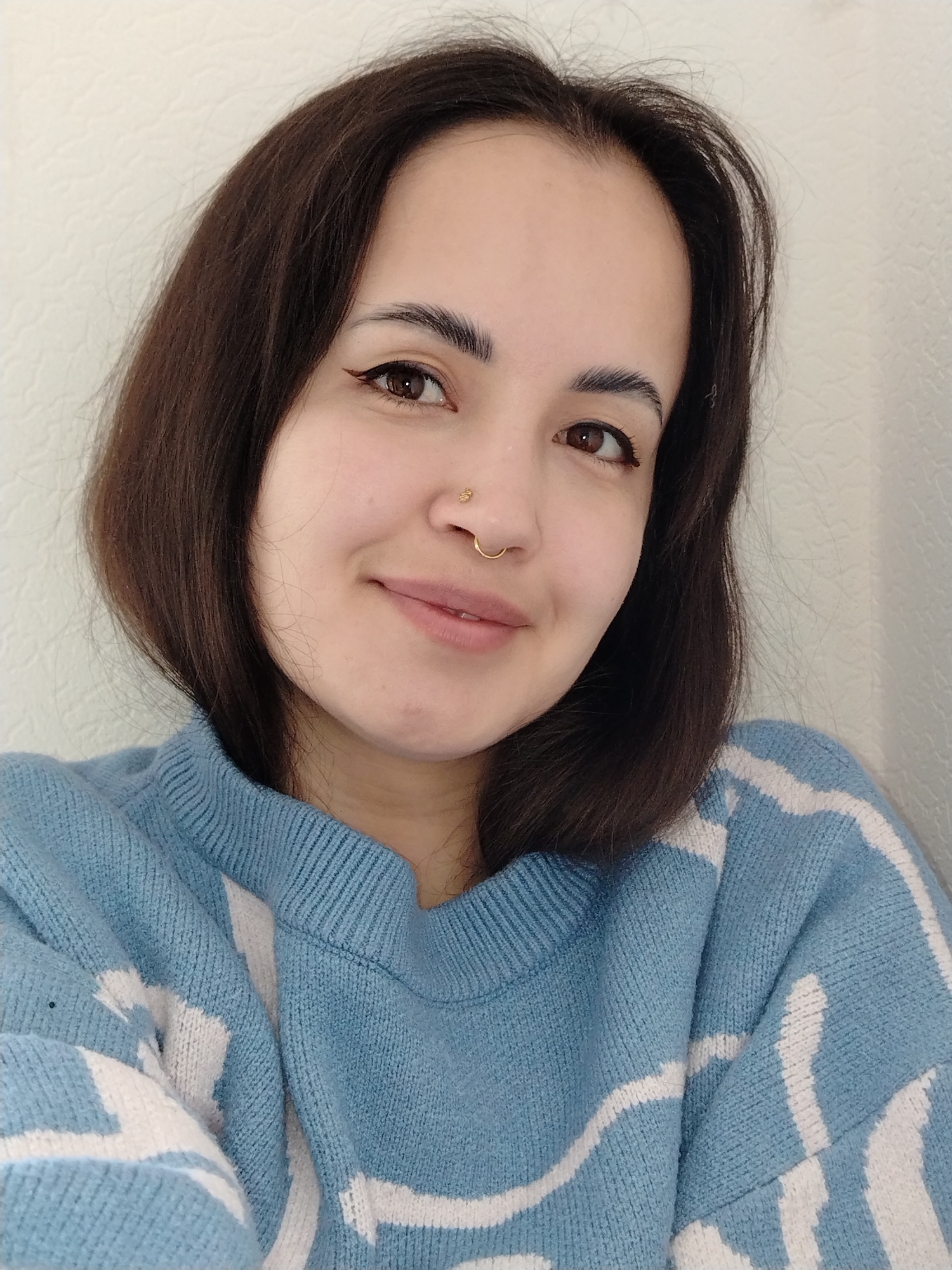 A close-up portrait features a young woman with a calm and gentle expression, captured in an indoor setting. She has medium brown, shoulder-length hair and warm brown eyes, accentuated by delicate eyeliner with a subtle wing. Her eyebrows are neatly shaped and full. She has two nose piercings: a small gold stud on her left nostril and a gold hoop on her right nostril. Her lips are gently closed in a soft smile, conveying a pleasant demeanor as she looks directly at the camera.
She is dressed in a light blue knitted sweater adorned with an abstract pattern of white, wave-like or angular stripes. The knit texture of the garment is clearly visible. The background consists of a plain, light-colored wall, likely off-white or beige, which has a subtle textured surface.
The lighting is soft and even, characteristic of an indoor environment during daytime. There are no specific weather indicators or visible text within the image. This scene is located in Noviy, Russia.