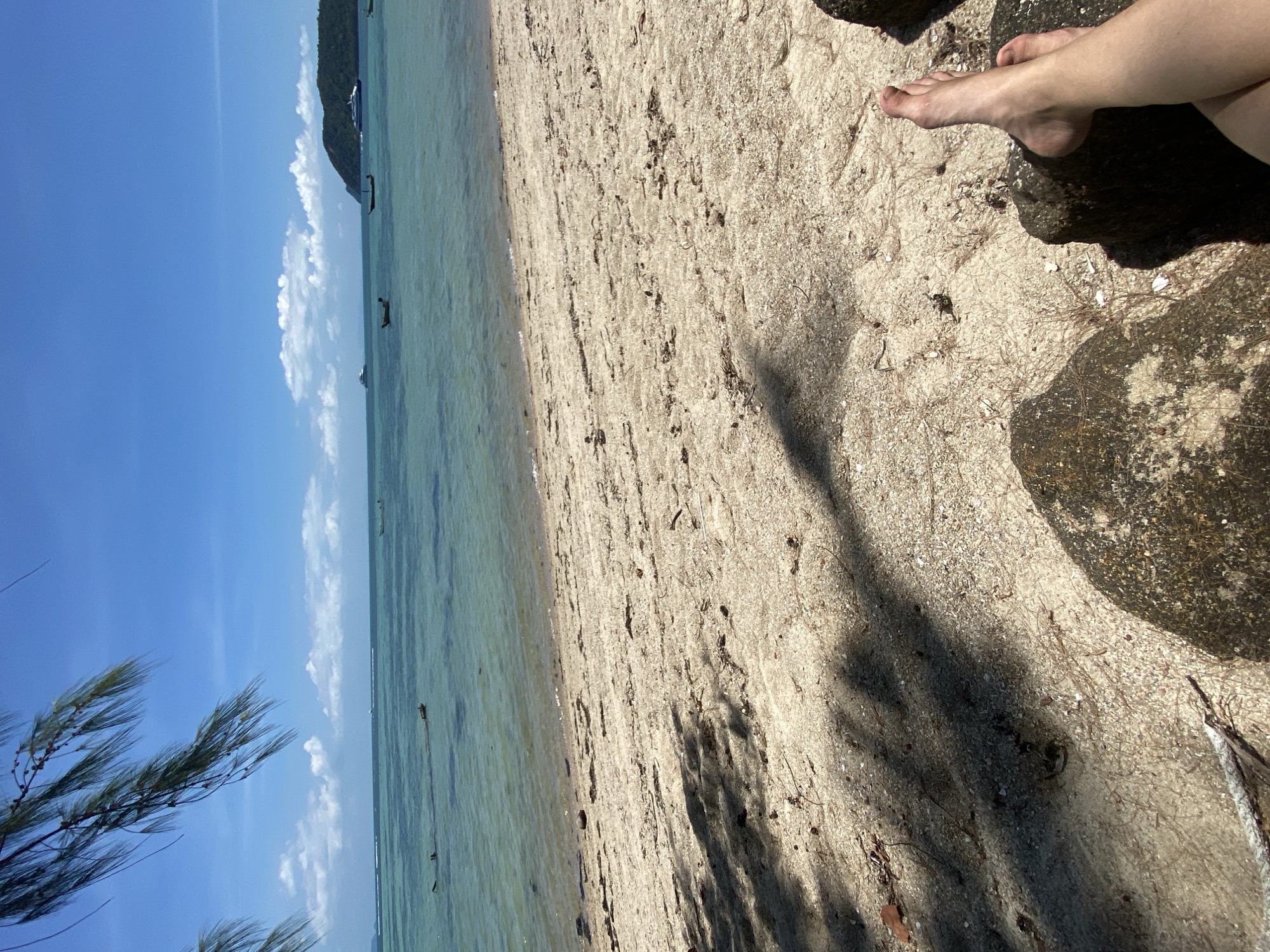 The image captures a serene beach scene, likely in Moo 1, Thailand, under bright daylight. The primary subjects are the natural landscape of a sandy beach meeting clear ocean waters, a bright blue sky, and a person's bare feet.
In the foreground, a sandy beach occupies the right half of the frame, showing fine, light-colored sand interspersed with small shells and dried plant debris, possibly pine needles. Large, dark, wet rocks are visible in the bottom right corner, partially in shadow. A pair of bare feet and lower legs of a person are visible in the top right, resting comfortably on or near the sand and rocks, suggesting relaxation. Strong, elongated shadows stretch across the sand from the upper right towards the center, indicating overhead sunlight, likely midday or early afternoon.
The midground transitions into calm, translucent blue-green ocean water that gently meets the shore. The water appears shallow near the beach, revealing the sandy bottom. In the background, the vast expanse of the ocean continues, dotted with several small boats, including what appear to be traditional long-tail boats and a larger vessel, hinting at local maritime activity. A distant, dark green, mountainous landmass or island is visible on the horizon to the top right.
The sky above is a vibrant, clear blue with a few scattered white cumulus clouds. In the top left corner, the feathery green foliage of what resembles a casuarina or pine tree branch partially frames the sky, swaying slightly as if in a gentle breeze. The overall mood is peaceful and tranquil, characteristic of a tropical beach setting. There is no visible text in the image.
