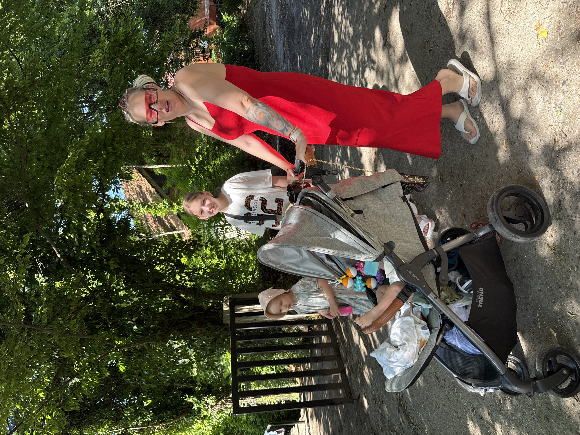 In Sochi, Russia, on a sunny day, a woman in a bright red dress and large red sunglasses stands beside a stroller. She has her right arm resting on the stroller's handle, and a colorful tattoo is visible on her left arm. Next to her and slightly behind, a young girl in a white t-shirt with a leopard print design looks towards the camera. A toddler in a light-colored dress and a hat is seated in the stroller, with their legs extended. The stroller is grey and has toys attached to it. In the background, a black metal gate and lush green trees are visible, suggesting a park or outdoor area. The ground is paved with a dirt and gravel mixture, dappled with sunlight and shadows.