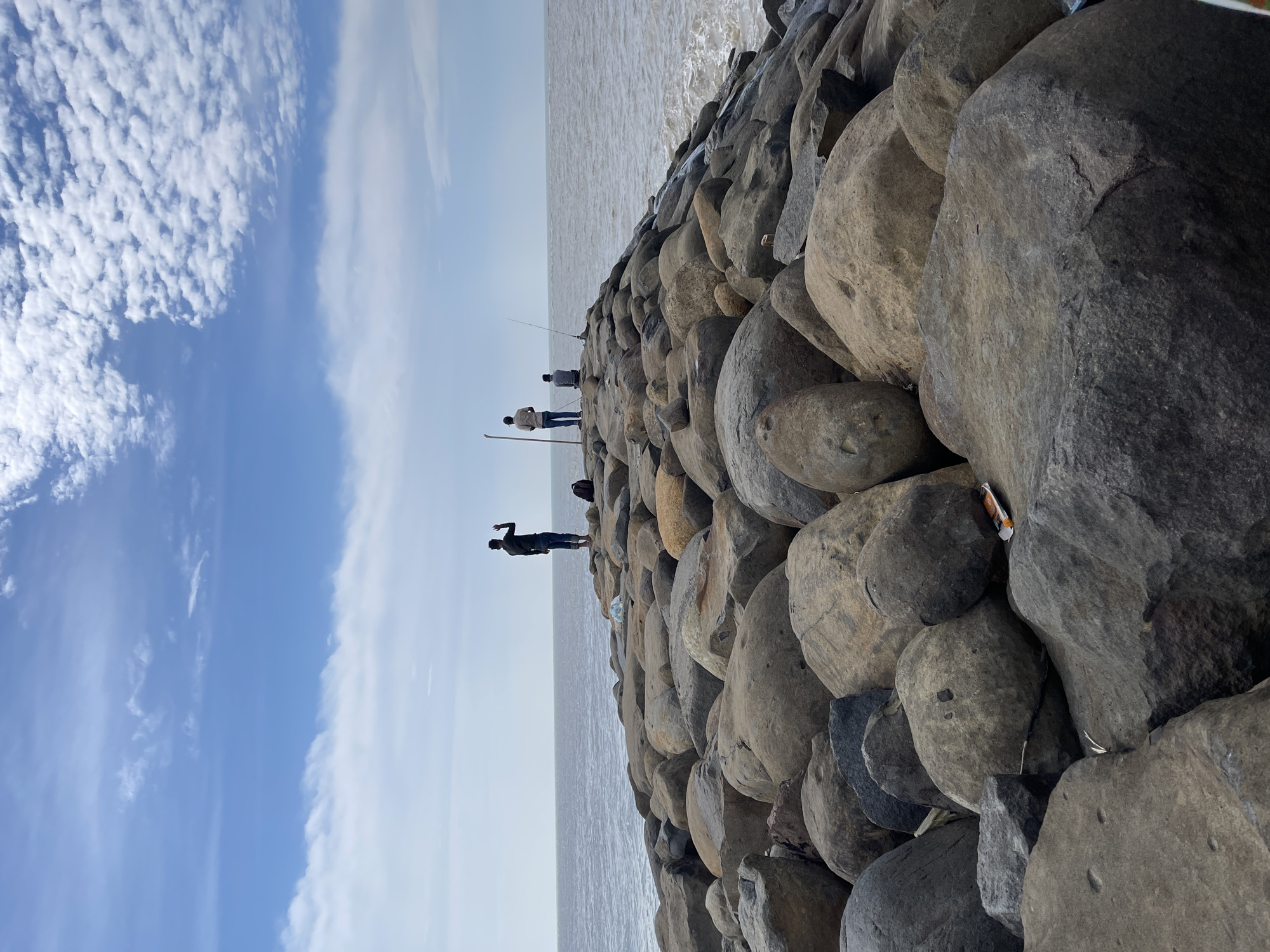 The image depicts a rocky pier extending into a body of water under a partly cloudy sky. In the distance, three individuals are visible on the pier. One person stands with their arm raised, seemingly waving or gesturing. Another individual is seated, holding a fishing rod, while a third person stands nearby, also with a fishing rod. The pier itself is constructed from large, rough-hewn rocks. The water appears calm with small waves lapping at the shore. The overall scene suggests a recreational fishing or leisure activity on a coastal area. The location is Kualacangkoi, Indonesia.