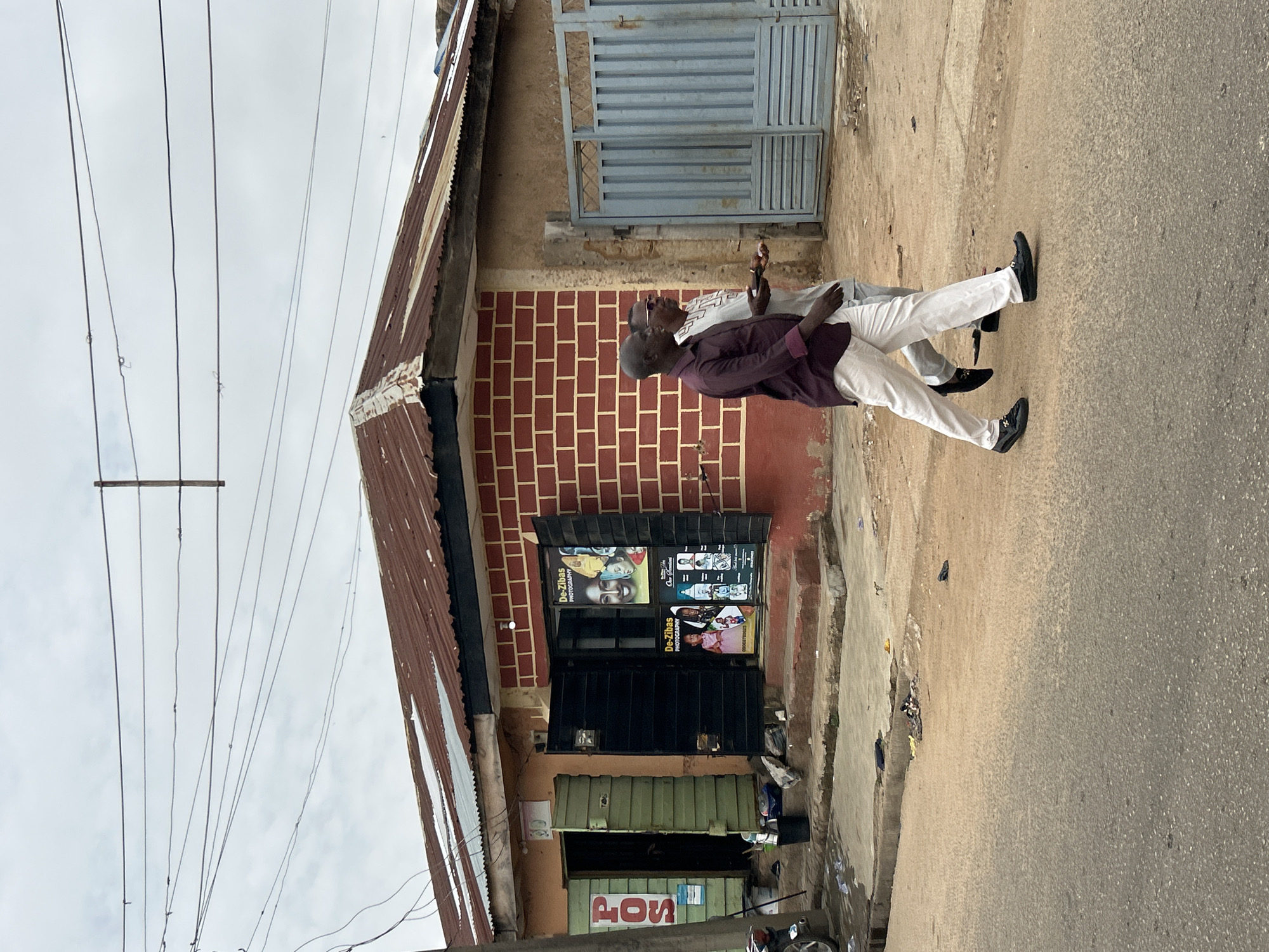 Two African men are walking past a building in Jos, Nigeria. One man is wearing a dark purple blazer, white pants, and black shoes. The other man is wearing a white shirt, white pants, and black shoes. The building is made of red bricks. The roof is corrugated metal. There are posters advertising photography services on the building. A gate is to the right of the brick building. There are power lines overhead.