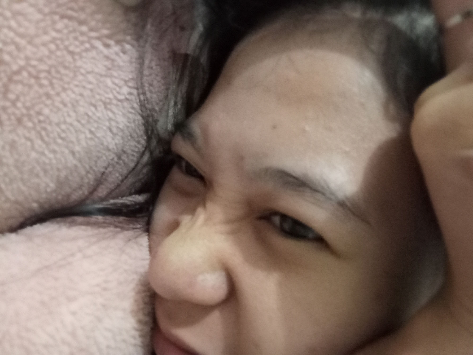 The image is a close-up, low-angle shot of a person's face, likely a child, lying down. The individual is positioned on their side, with their head resting on a pink, fuzzy surface, possibly a blanket or pillow. Their face is contorted in what appears to be a playful or perhaps slightly uncomfortable expression, with eyes squeezed shut and the nose wrinkled. Dark hair frames the face. A portion of an adult's hand is visible at the top of the frame, appearing to gently hold or cover the person's head. The lighting is soft, and the background is blurred, focusing attention on the facial expression. The scene is domestic and informal. There is no visible indication of location within Rejosari, Indonesia.