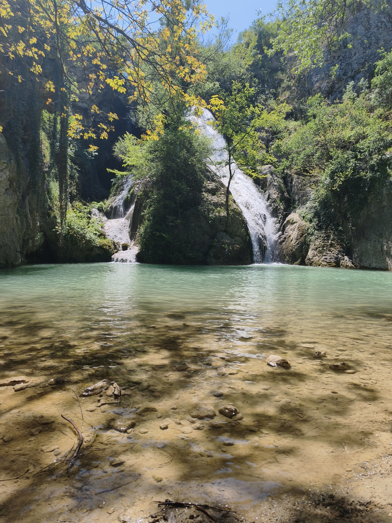 The image depicts a scenic waterfall and pool located in Hotnitsa, Bulgaria. The waterfall cascades down a rocky cliff face into a clear, turquoise-colored pool. Lush vegetation covers the surrounding cliffs, with trees and vines visible. The foreground shows a shallow, sandy area with rocks and tree debris, indicating a natural, undisturbed environment. Sunlight filters through the trees, creating shadows and highlighting the clarity of the water. No people are visible in the scene.