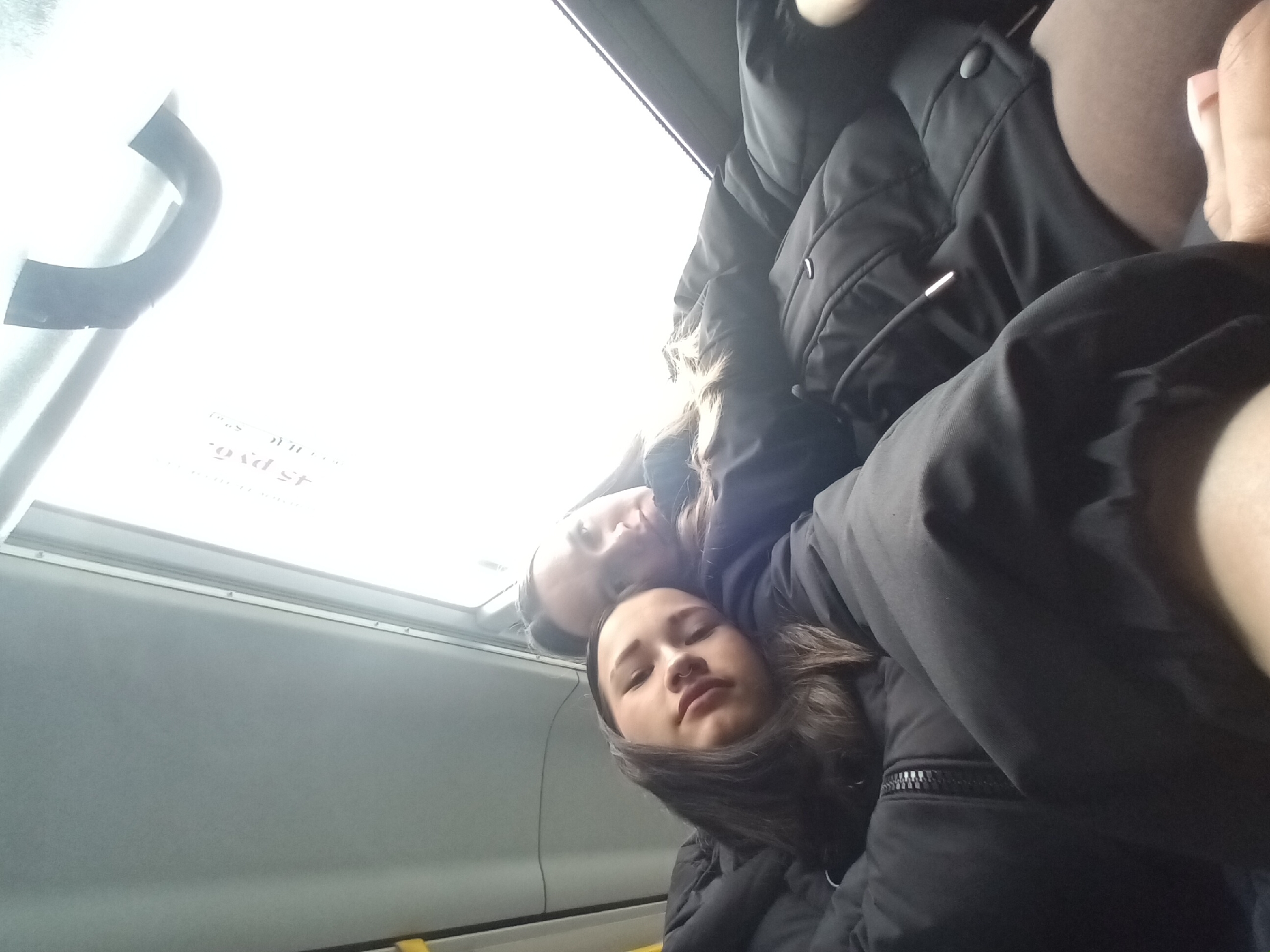 Two young women are close together, appearing to be on a bus or train. They are wearing dark winter jackets. The woman in the foreground has dark hair and is looking at the camera. The other woman is lying behind her, also with her eyes closed. The interior of the bus or train is visible, including a window and a handrail. The exact location within the city of Chaikovsky, Russia cannot be determined from the image alone.
