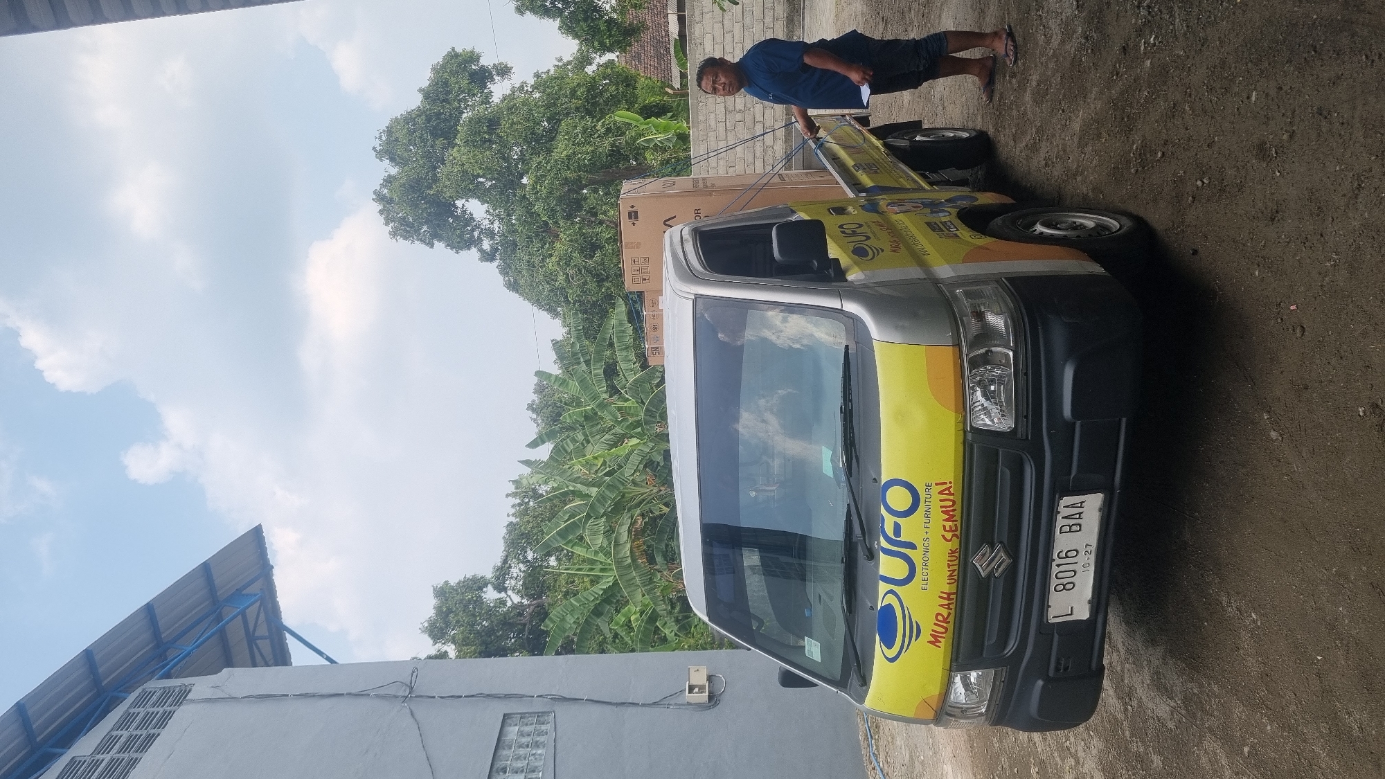 A man stands next to a silver Suzuki Carry pickup truck parked on a dirt road in front of a building. The truck has a yellow graphic on the side with "UFO ELECTRONICS + FURNITURE MURAH UNTUK SEMUA!" text and a blue UFO logo. Two large cardboard boxes are secured with ropes in the truck bed. The man is wearing a dark blue t-shirt, dark shorts, and flip-flops. He is holding a piece of paper. The building in the background is grey with a blue awning and a metal grate. Banana trees and other greenery are visible behind the truck. The sky is partly cloudy. The license plate on the truck reads "L 8016 BAA". The image was taken in Diwek, Indonesia.