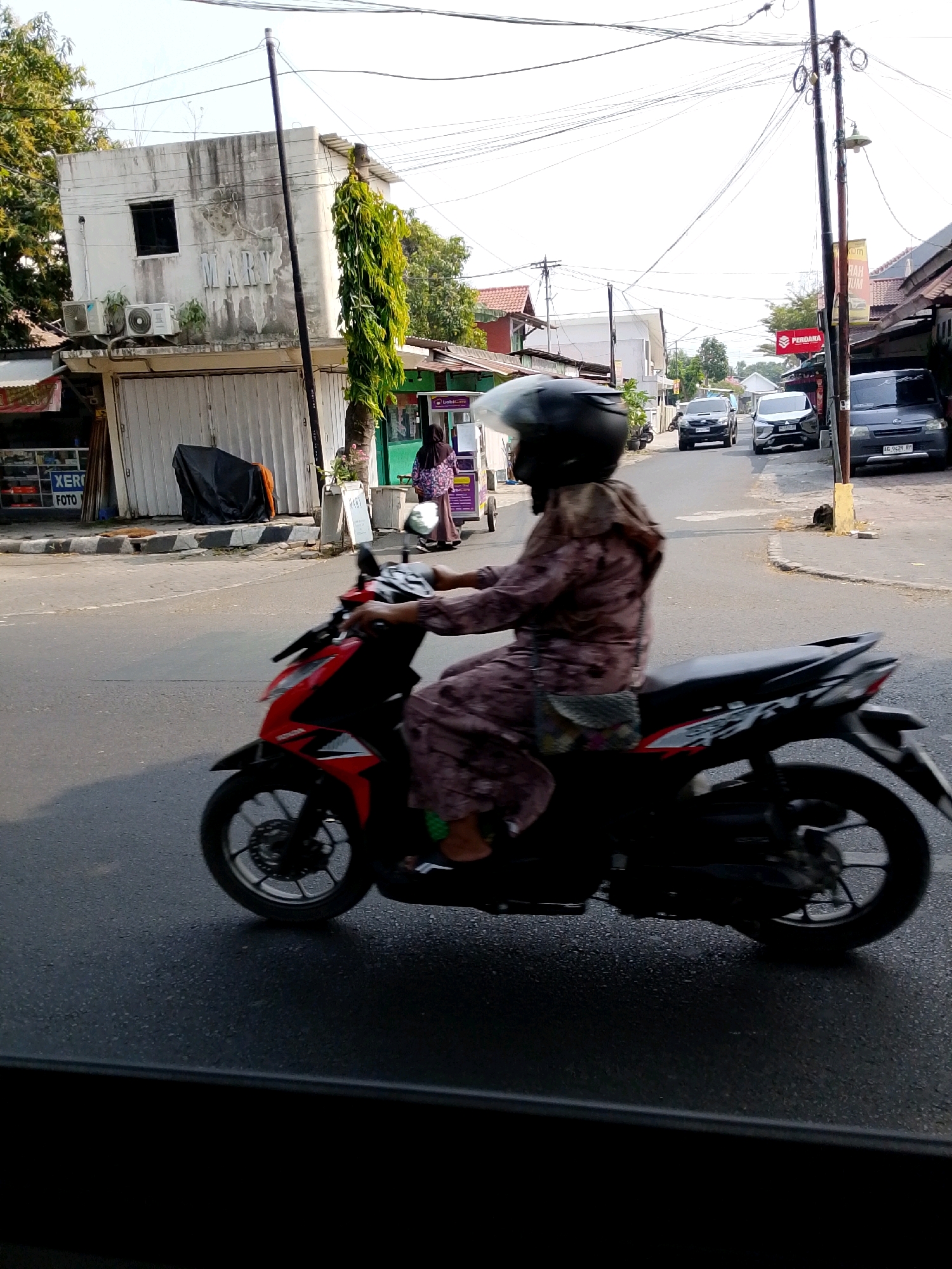 A side-view, medium shot captures a person riding a red and black motorcycle down a paved road in Kediri City, Indonesia. The rider, wearing a black helmet and a patterned long-sleeved shirt with a scarf, is partially obscured by the motion blur of the motorcycle. The motorcycle is in motion, with its wheels and body blurred.
In the background, the environment suggests a street scene with several buildings. To the left, a white building with a black-framed window and a sign that reads "MARY" is visible, along with two air conditioning units. A vertical green tree stands beside it. Further down the street, other buildings and a red sign with white lettering are present. Several cars are parked or driving on the road in the distance. The sky is bright and overcast. The road surface appears wet in places, reflecting the ambient light. The foreground is dominated by the motorcycle and the wet asphalt.