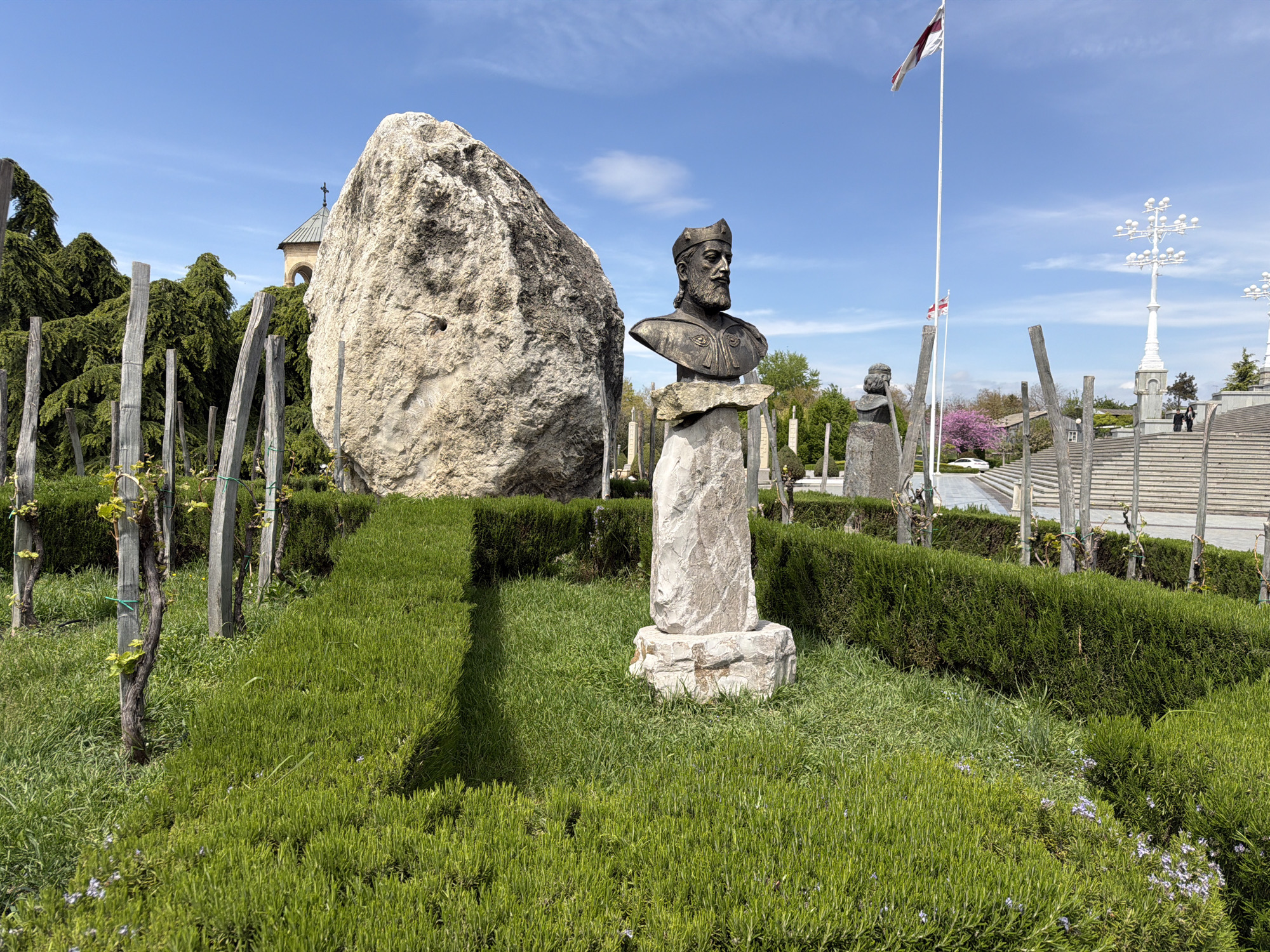 This daytime image captures a serene outdoor scene, likely a memorial park or garden, in Tbilisi, Georgia, under a clear blue sky with wispy clouds. The foreground and midground are dominated by lush green grass and neatly trimmed dark green hedges forming pathways and garden beds. Rows of young grapevines on wooden posts are visible throughout the garden.
The central feature is a bronze bust of a bearded man wearing a crown or ornate headpiece, set atop a rough stone pedestal. To its immediate left stands a large, light-colored natural boulder. In the background, to the right, other similar busts or statues can be seen.
Further back on the right, a wide stone staircase ascends. Two people are visible walking up these stairs. Tall white flagpoles fly the Georgian flag (a red cross on a white background), and decorative white multi-branched streetlights illuminate the area around the staircase. A white car is partially visible near the bottom of the stairs, and a tree with delicate pink blossoms adds a splash of color. In the upper left, behind a canopy of dense evergreen trees, a building with a conical roof and a cross on its spire suggests a church or monastery. The overall setting implies a site of historical and cultural significance, possibly celebrating Georgian heritage or its renowned wine culture.