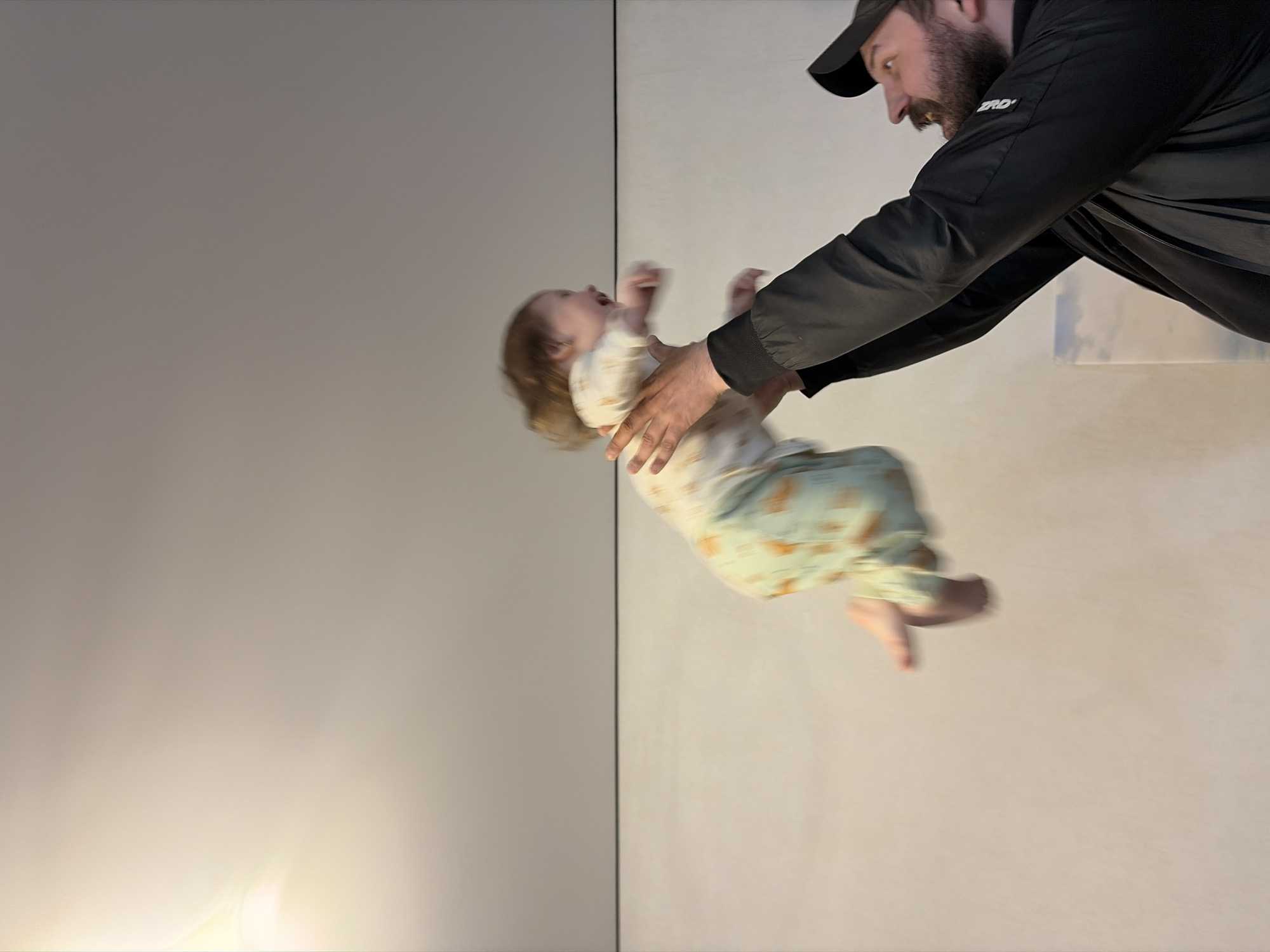 A man in a black jacket and cap is holding a baby in the air. They are indoors, against a neutral-colored wall. The image is taken from an unusual angle, making it appear as if the subjects are floating. No information about the location is discernible.