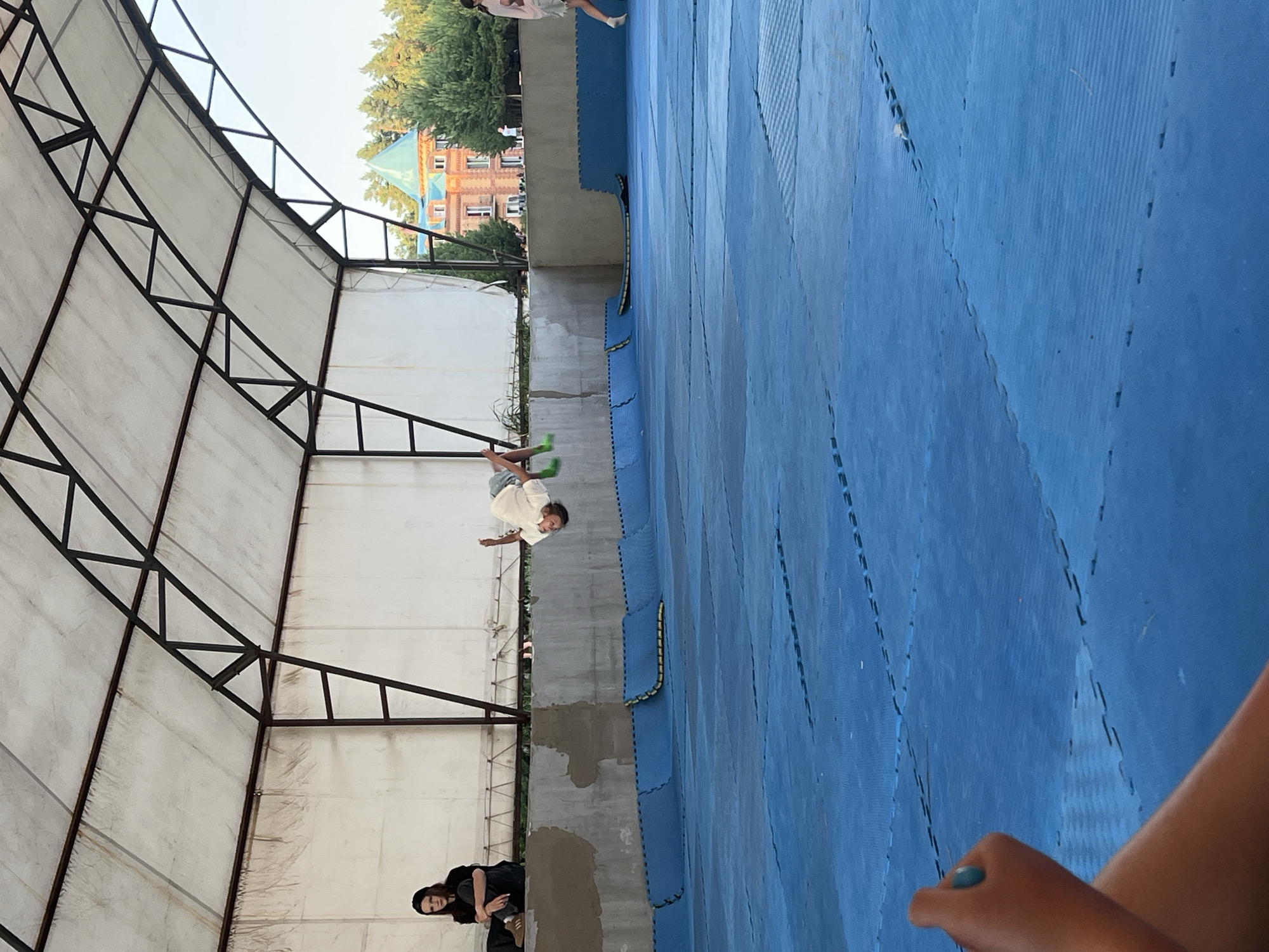 The image depicts an indoor gymnasium with blue matting on the floor and a white, tent-like roof supported by a metal frame. In the center of the frame, a young person in a white t-shirt and grey shorts is captured mid-air performing a gymnastic flip. To the left, a young woman wearing a black hijab and dark clothing sits on a raised concrete ledge. In the background, a red brick building with a green turret is visible amongst trees. The foreground shows a portion of a person's arm and leg, suggesting the photographer's perspective. The scene is likely a recreational or training facility.