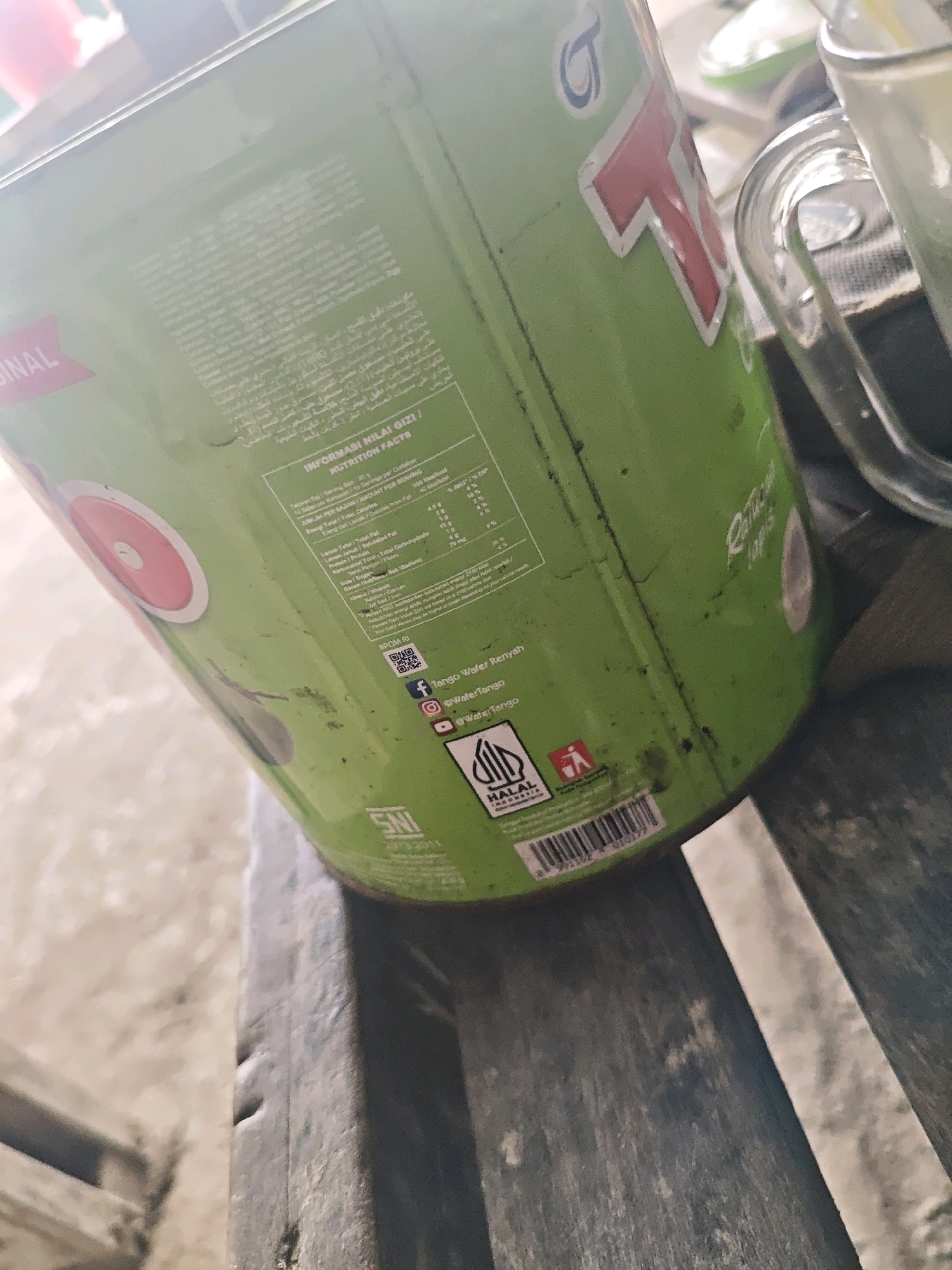 The image shows a close-up of a green cylindrical container, likely packaging for a food product, sitting on a wooden surface. The container has visible text and logos, including what appears to be "TANGO" in large red lettering. A portion of a glass with a straw is visible to the right of the container. The background is blurred but suggests an outdoor setting, possibly a table with a textured surface. The lighting indicates it is daytime. No specific location in Kemuning, Indonesia is discernible from the image.