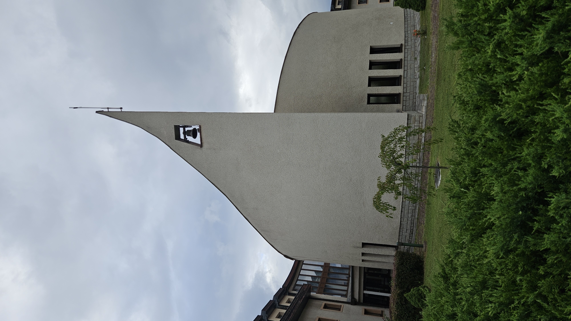A contemporary building with a large, curved facade dominates the frame, its rough, light-colored plaster contrasting with the overcast sky. A sharply angled protrusion rises from the facade, housing a bell within a square opening. A slender cross, attached to a metal pole, tops the angled structure. The building's base is lined with a row of tall, narrow windows. To the left, a portion of an adjacent building with a more traditional sloped roof and dormer windows is visible, featuring large glass panels. In the foreground, a dense, green hedge stretches across the bottom of the image. Behind the hedge, a patch of manicured lawn is visible, with a small, weeping tree planted centrally and a single, small orange flower near the building's base. The setting appears to be a modern church or religious institution, possibly in Prešov, Slovakia, given the architectural style. There are no people or animals visible in the image.