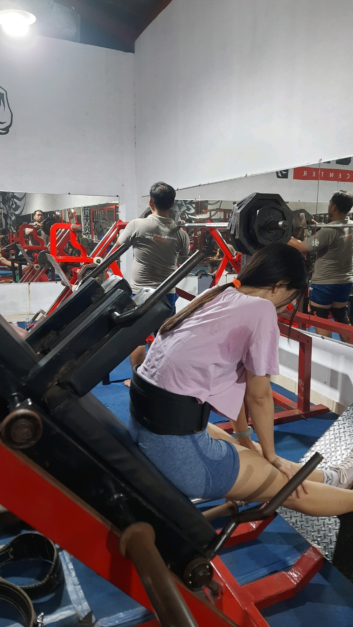 In a gym, a woman wearing a pink t-shirt and blue shorts is seated on a leg press machine, facing away from the camera. She is wearing a weightlifting belt. Her hair is pulled back in a ponytail. In the background, two men are also engaged in weightlifting. One man is wearing a grey t-shirt and is seen from the back as he lifts weights on a rack. The other man, also wearing a grey t-shirt, is further in the background, facing a mirror. The gym has white walls and blue mats on the floor. Mirrors line one wall, reflecting the gym equipment and patrons. There is a large weight plate visible on a barbell.