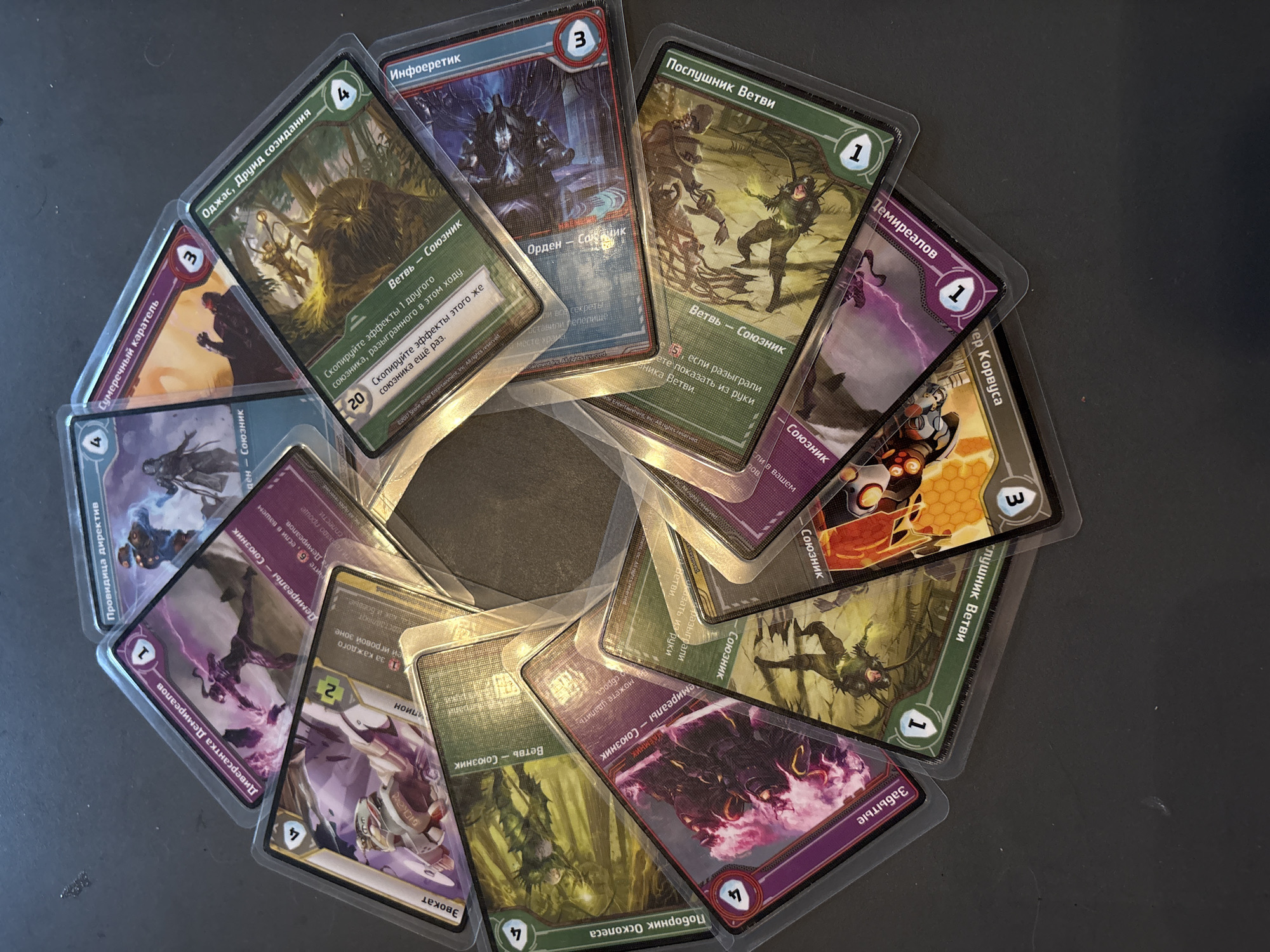 The image displays a circular arrangement of numerous playing cards, fanned out and overlapping each other on a dark, textured surface. The cards feature colorful illustrations and text in Cyrillic script. Some cards are upright, while others are tilted at various angles, creating a dynamic composition. The lighting suggests a bright, overhead source, casting subtle shadows. There are no people visible in the image. The background is a plain, dark color, likely a table or floor. This image is not located in Shymkent, Kazakhstan; it is a general still life of playing cards.